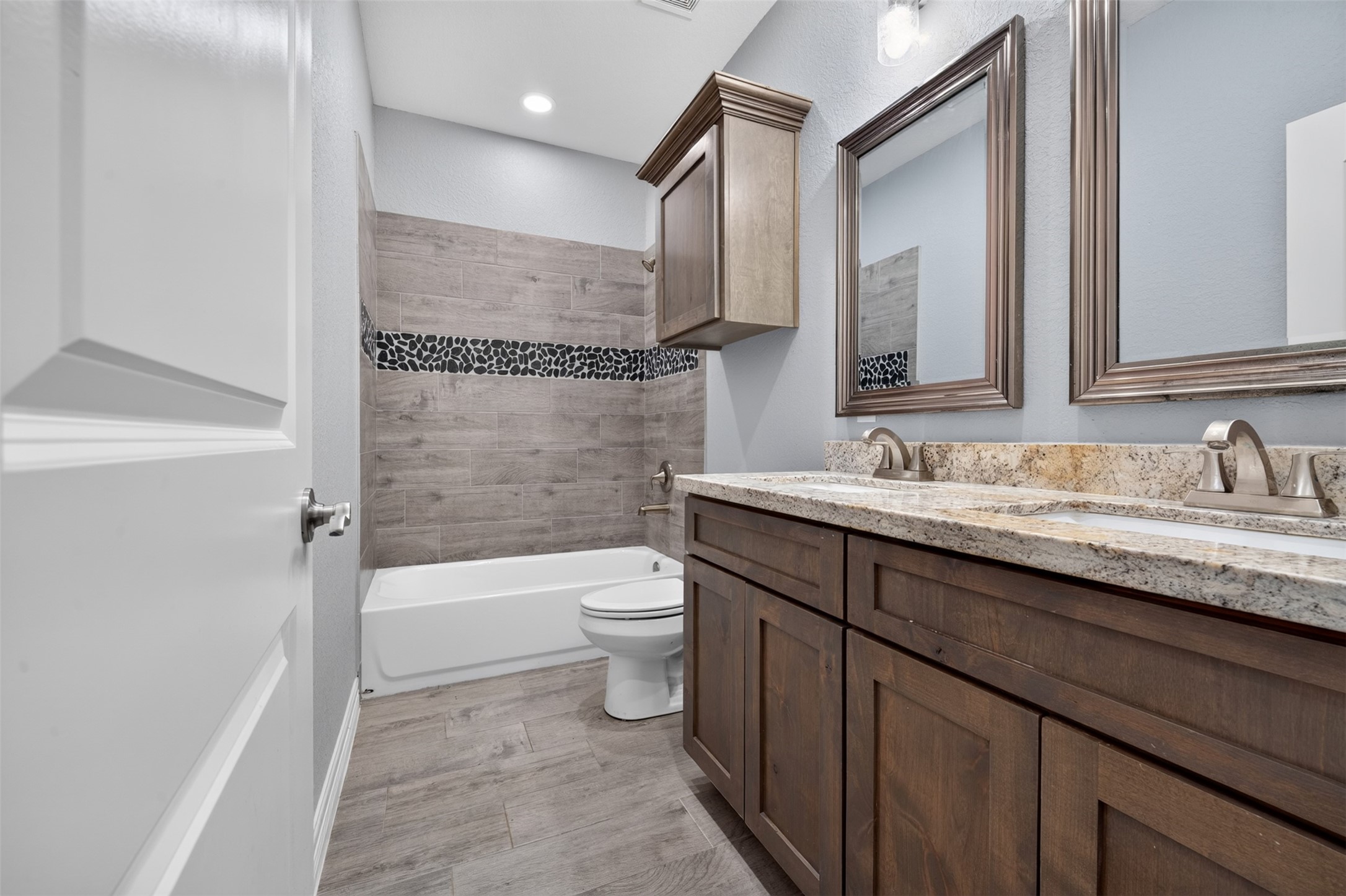 Elegant bathroom featuring dual sinks with granite countertops, rich wooden cabinetry, and a stylish tiled shower with a pebble accent strip.