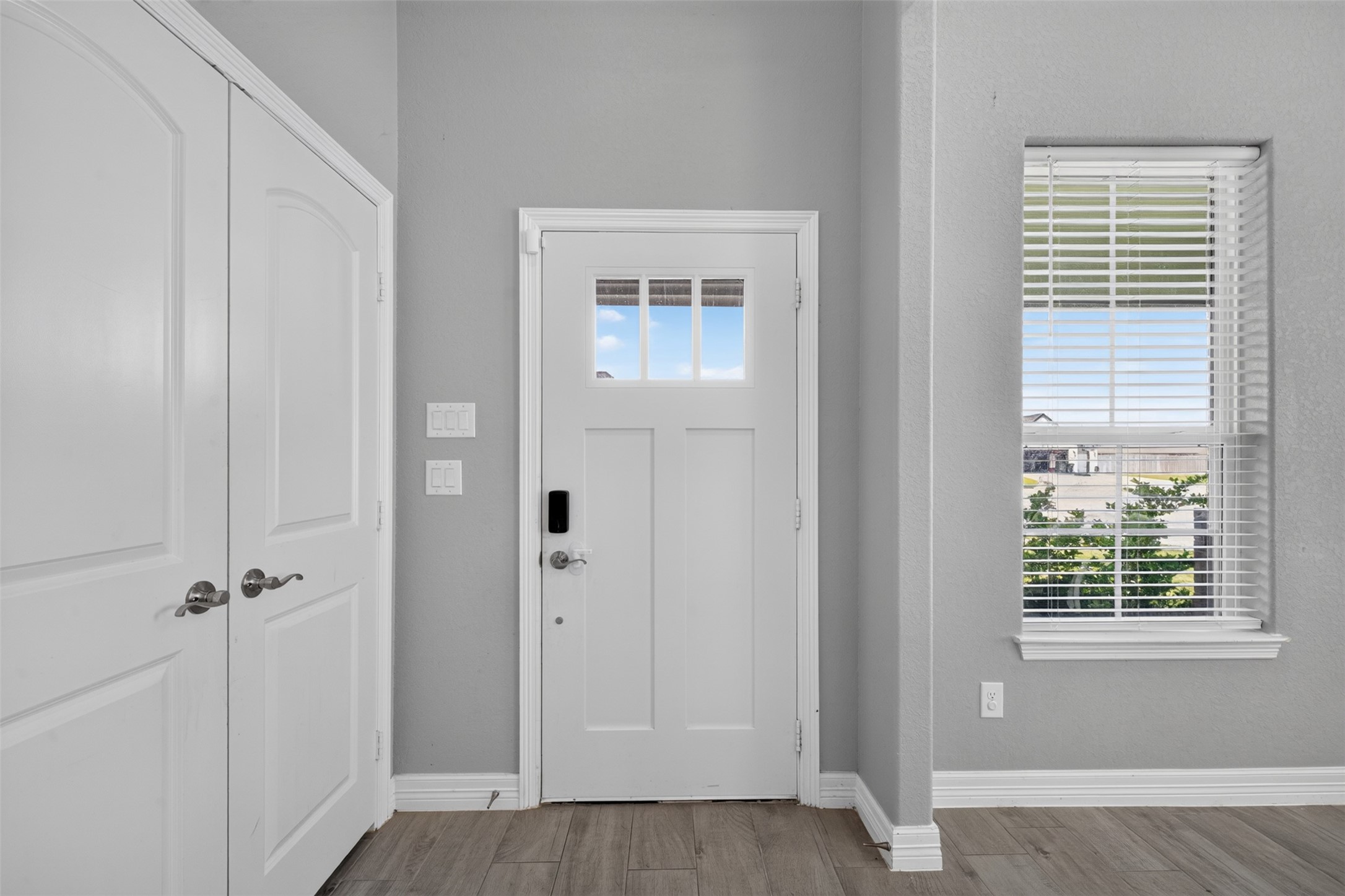 Inviting entryway features a sleek front door with a window panel, flanked by a bright window and ample closet space, all accented by modern gray walls and wood-look flooring.