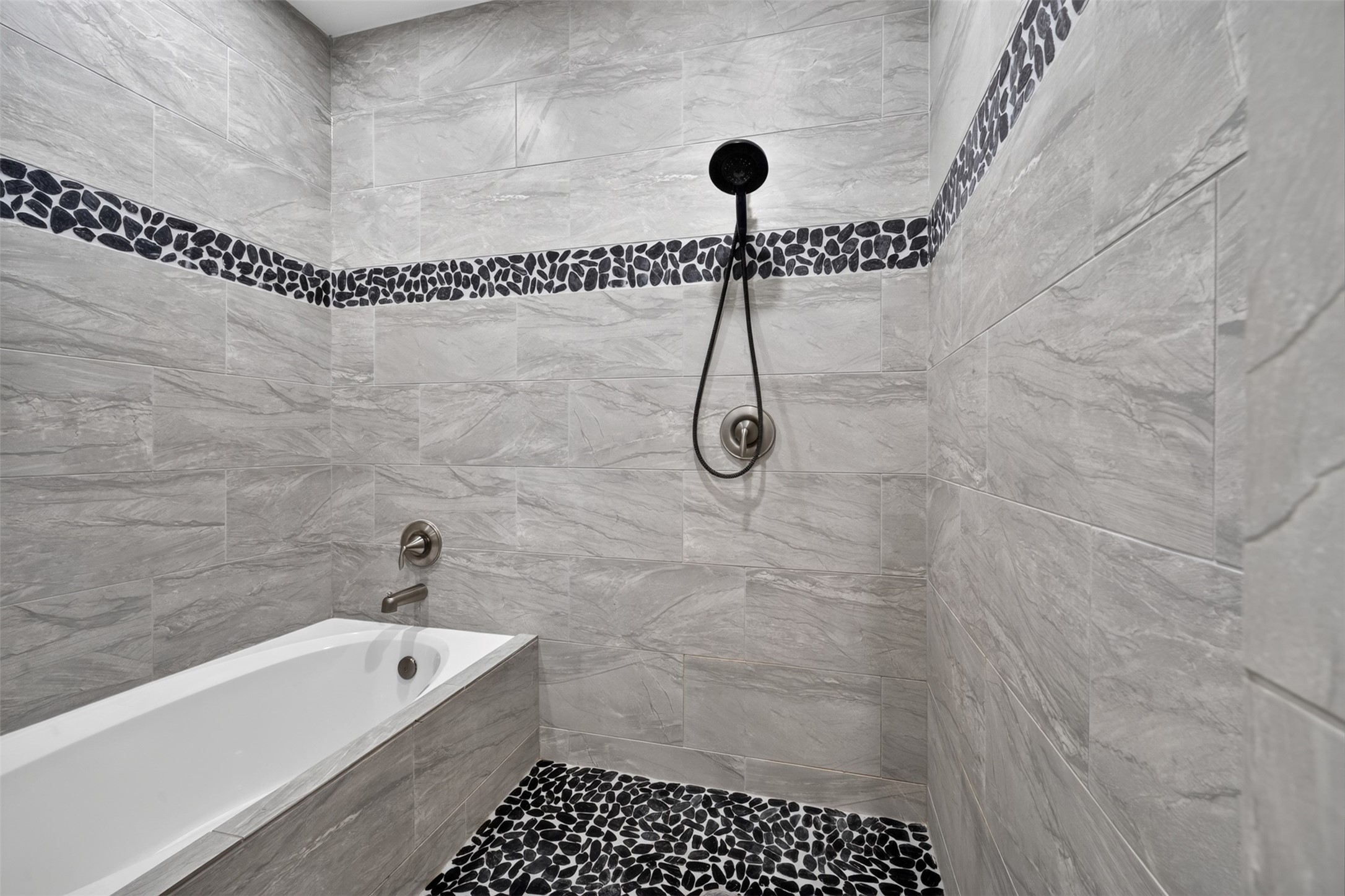 Elegant bathroom featuring a luxurious tub, sleek gray tiles with a stylish pebble accent strip, and a modern handheld showerhead for a spa-like experience.