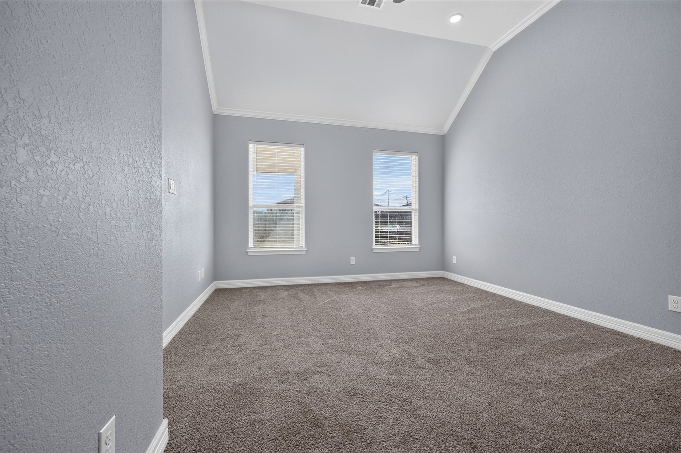 Spacious room with high vaulted ceilings, soft gray walls, and plush carpeting, featuring two large windows that invite natural light.