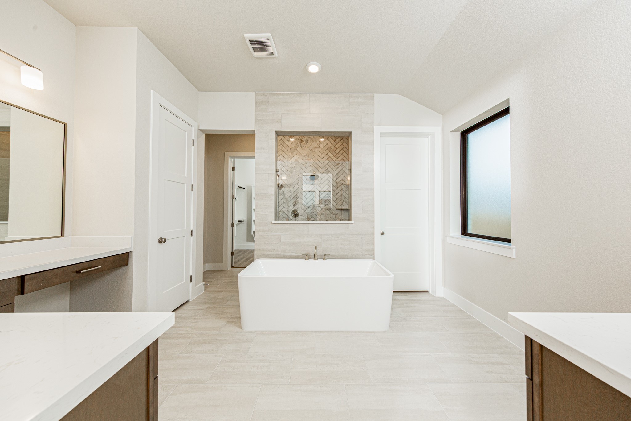 This elegant primary bathroom features a stylish dual-vanity layout with stained wood cabinetry, sleek brushed nickel fixtures, and clean-lined framed mirrors. Bright quartz countertops and modern lighting create a fresh, upscale feel, while the space opens seamlessly to the primary suite beyond, enhancing both flow and functionality.