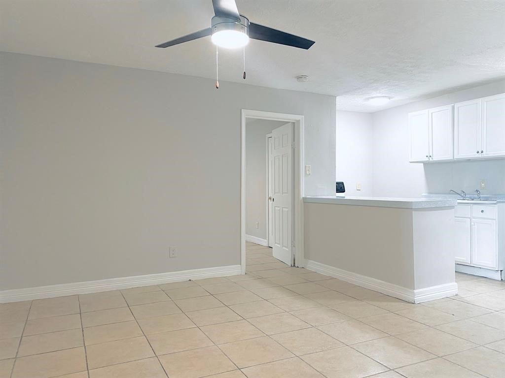 Spacious open-concept living area with neutral tiles, a modern ceiling fan, and a bright kitchen featuring white cabinetry.