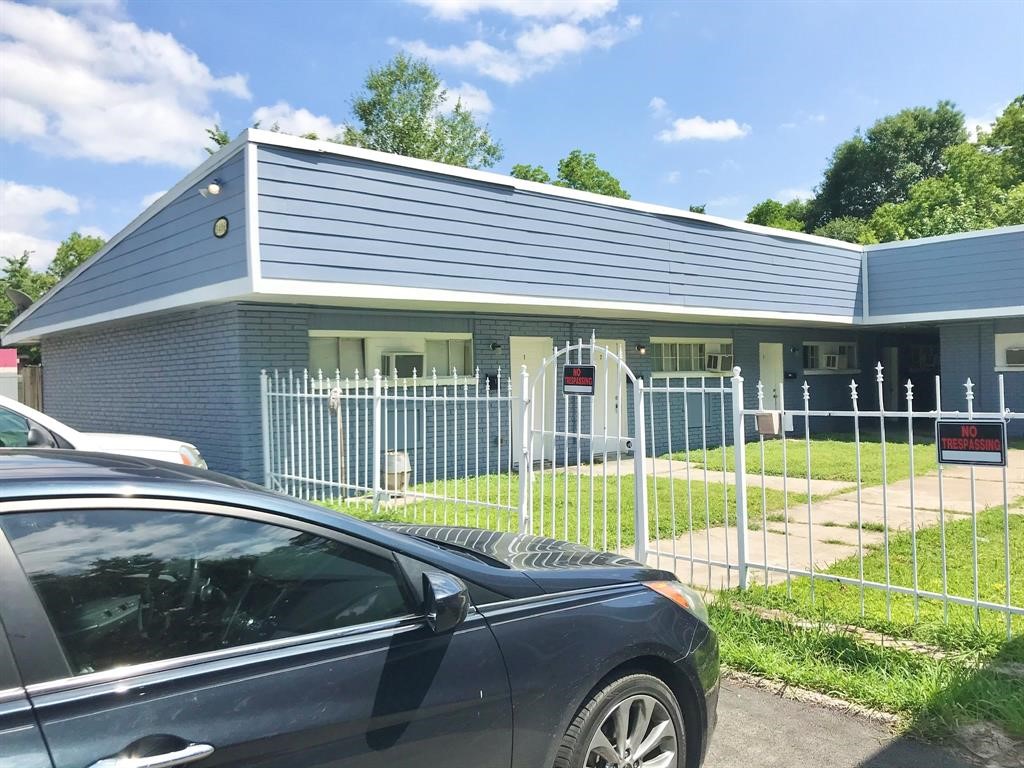 Charming blue brick home with a modern flat roof, surrounded by a secure white fence, offering a neat front yard and convenient driveway access.