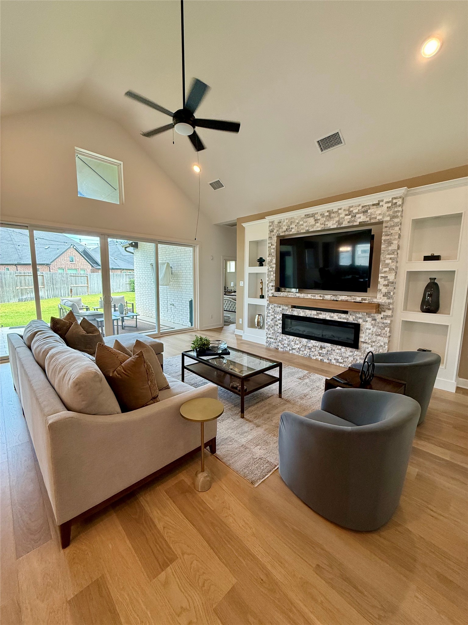 Spacious living room with high ceilings, elegant wood flooring, and custom designed modern stone accent wall featuring a built-in fireplace and TV. Large windows provide ample natural light and offer views of the backyard.