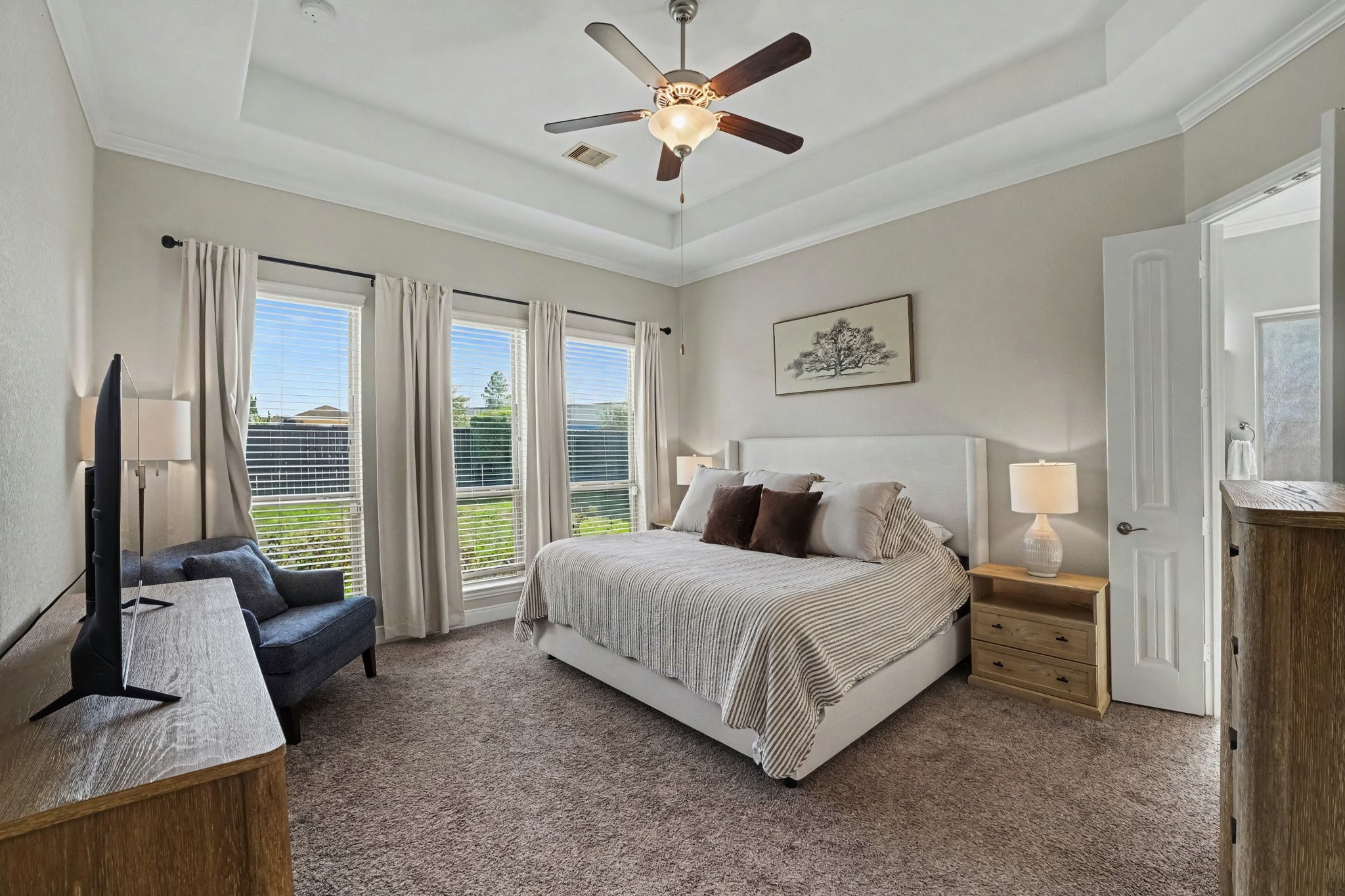 Three large floor-to-ceiling windows equipped with privacy blinds and flowing neutral drapes flood the bedroom with soft daylight while providing pleasant views of the lush backyard.