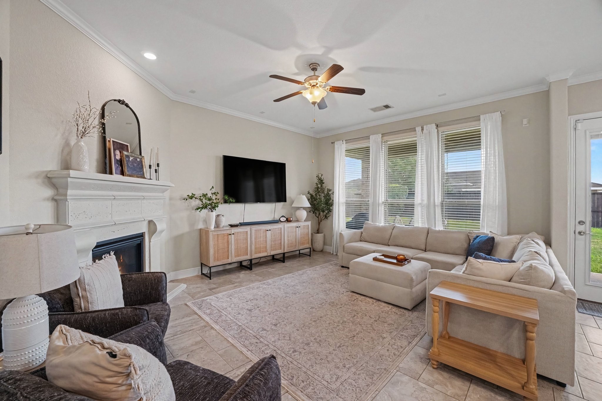 This bright and spacious living room serves as the heart of the home, offering an inviting space designed for both relaxation and entertaining. High ceilings with crown molding, recessed lighting, and a classical wooden ceiling fan set a airy, elevated tone. Wall-to-wall windows topped with sheer drapes flood the room with natural light while providing views out to the backyard.