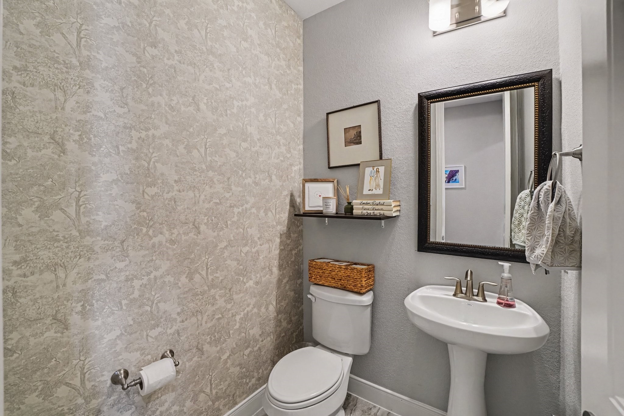 This charming powder room offers a stylish and convenient space for visiting guests. One accent wall is dressed in a subtle, elegant tree-patterned wallpaper, adding personality and texture to the room. A classic white pedestal sink with brushed nickel hardware pairs with a dark-framed rectangular mirror and warm overhead lighting.