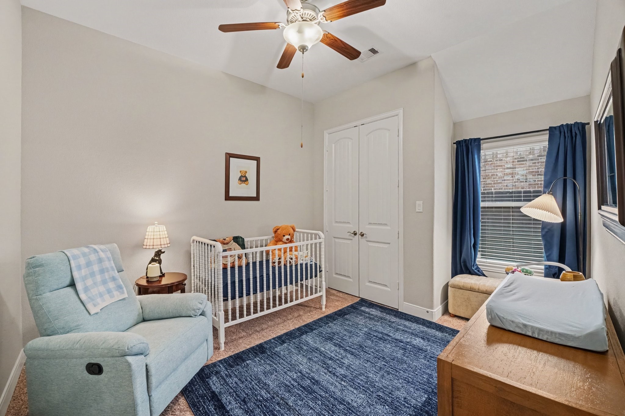 This inviting secondary bedroom, currently outfitted as a cozy nursery, features vaulted ceilings that add an airy and spacious feel to the room. A classic wooden ceiling fan with an integrated light fixture anchors the top of the space, while neutral grey walls and plush carpeting create a comfortable environment underfoot.