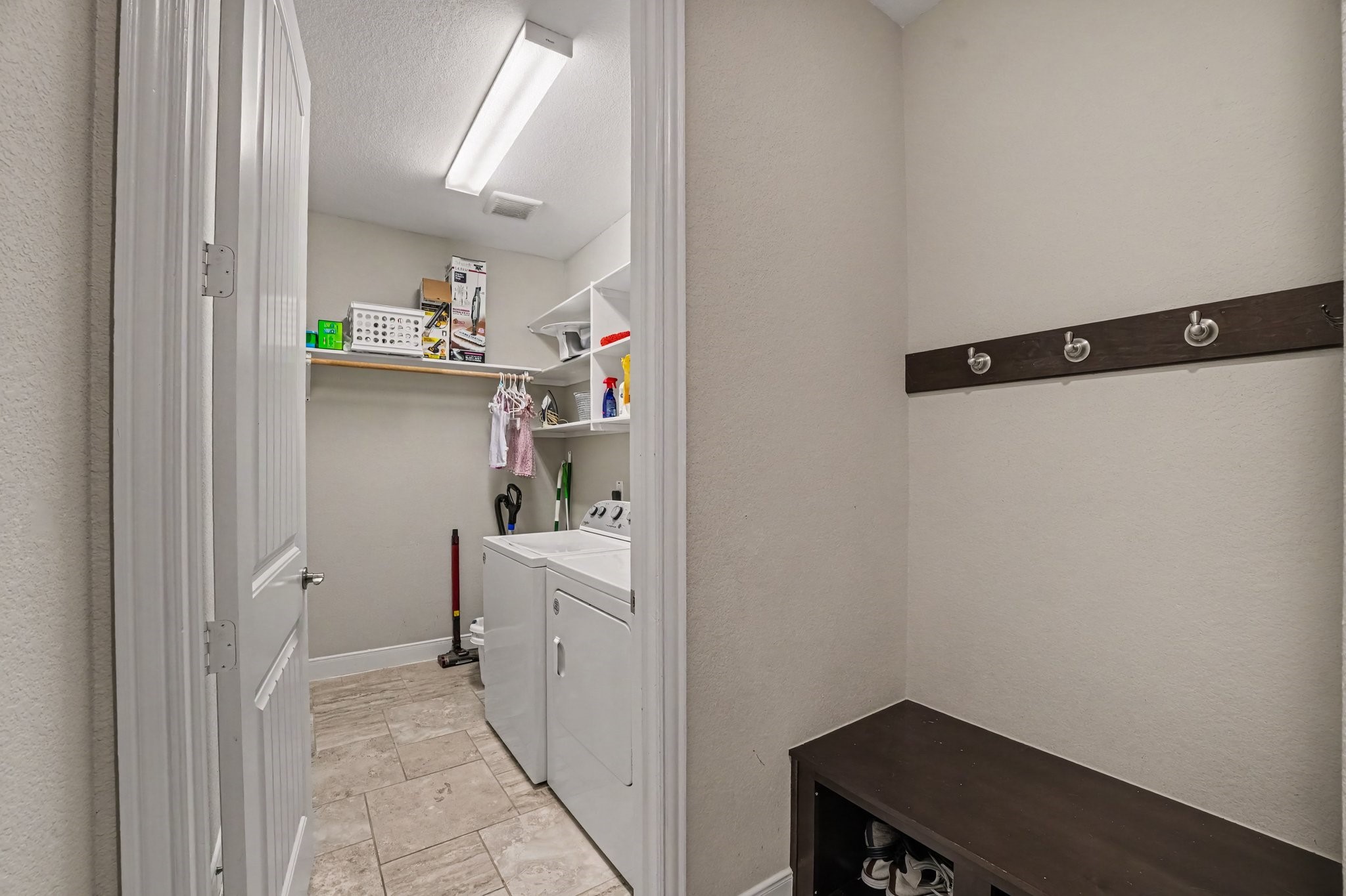 This dedicated utility hub highlights everyday convenience with a functional laundry room and mudroom entry drop zone. Right outside the laundry door, a dark wood bench with shoe storage cubbies and a wall-mounted hook rail provides the perfect spot to kick off shoes and hang coats or bags. Stepping through the doorway reveals a spacious laundry room equipped with a side-by-side washer and dryer.
