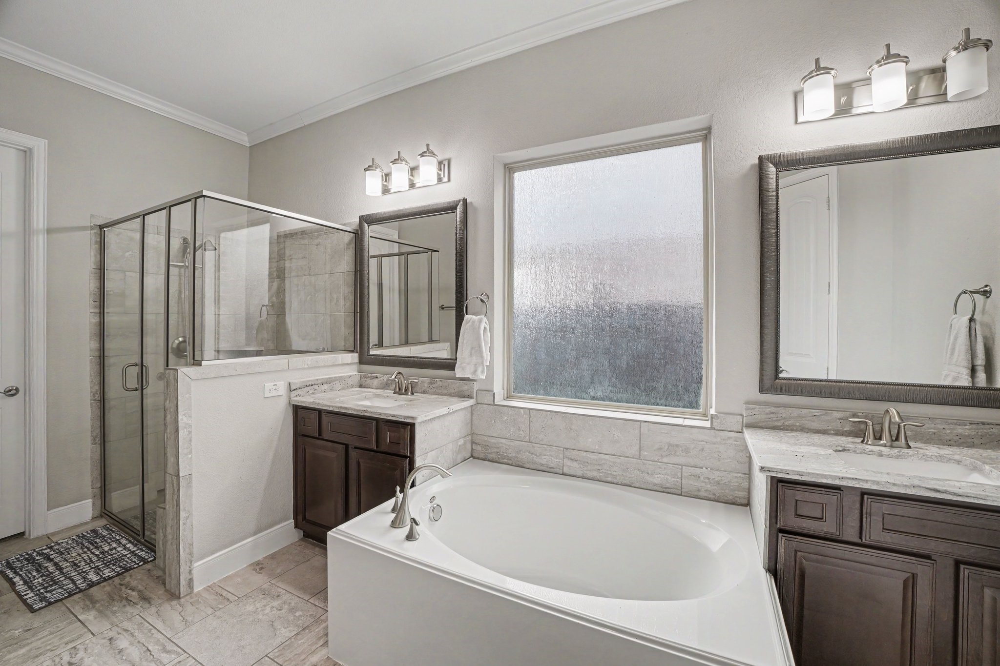 At the heart of the space lies a deep, oval soaking tub nestled beneath a large frosted privacy window, filling the room with soft natural light while maintaining complete seclusion. Flanking the tub are separate dual vanities featuring rich dark cabinetry, sleek granite countertops, brushed nickel hardware, framed mirrors, and contemporary three-light vanity fixtures.