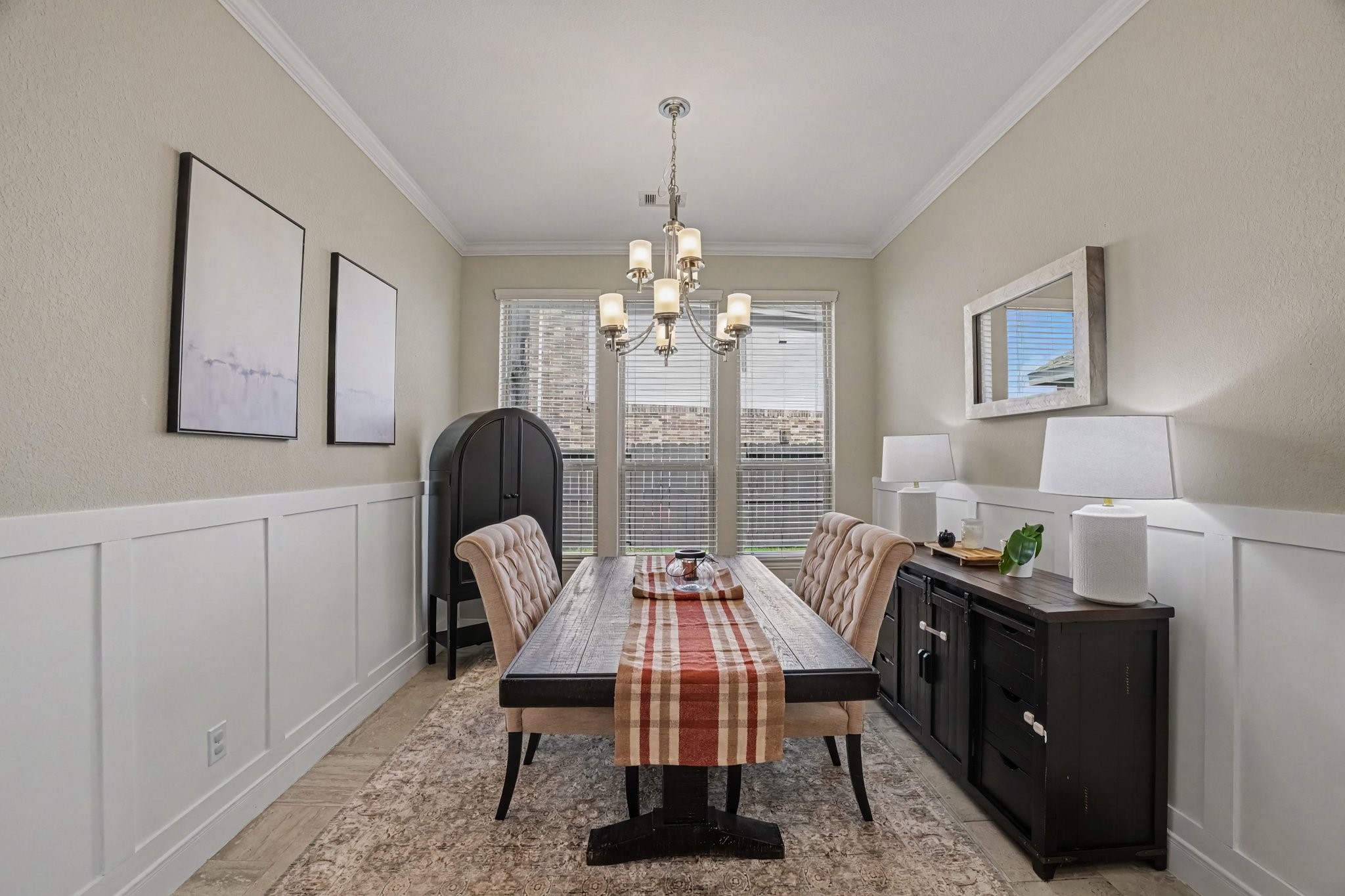 This sophisticated formal dining room exudes elegance and charm, making it the perfect venue for intimate family dinners and festive gatherings. Crisp white board-and-batten wainscoting grounds the space with classic architectural detail, while refined crown molding frames the soft, neutral walls above. At the center of the room hangs an eye-catching, traditional chandelier that illuminates a rich, dark-wood dining table paired with stylish tufted upholstered chairs.