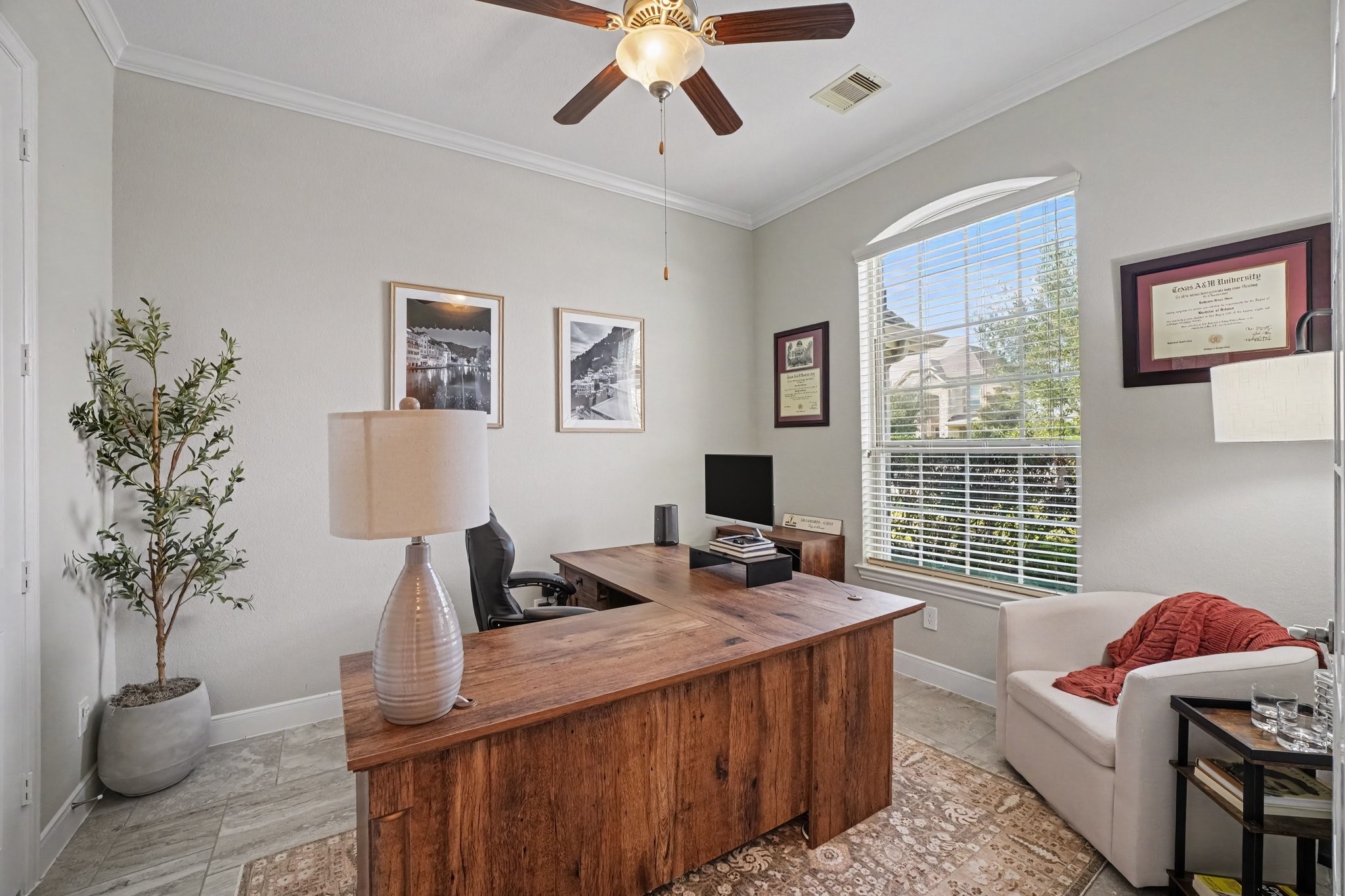 This dedicated home office offers an exceptional space for remote work, study, or management. High ceilings with crisp crown molding, a stylish wooden ceiling fan with integrated lighting, continuous tile flooring, and a decorative potted olive tree bring polish and warmth to the space.