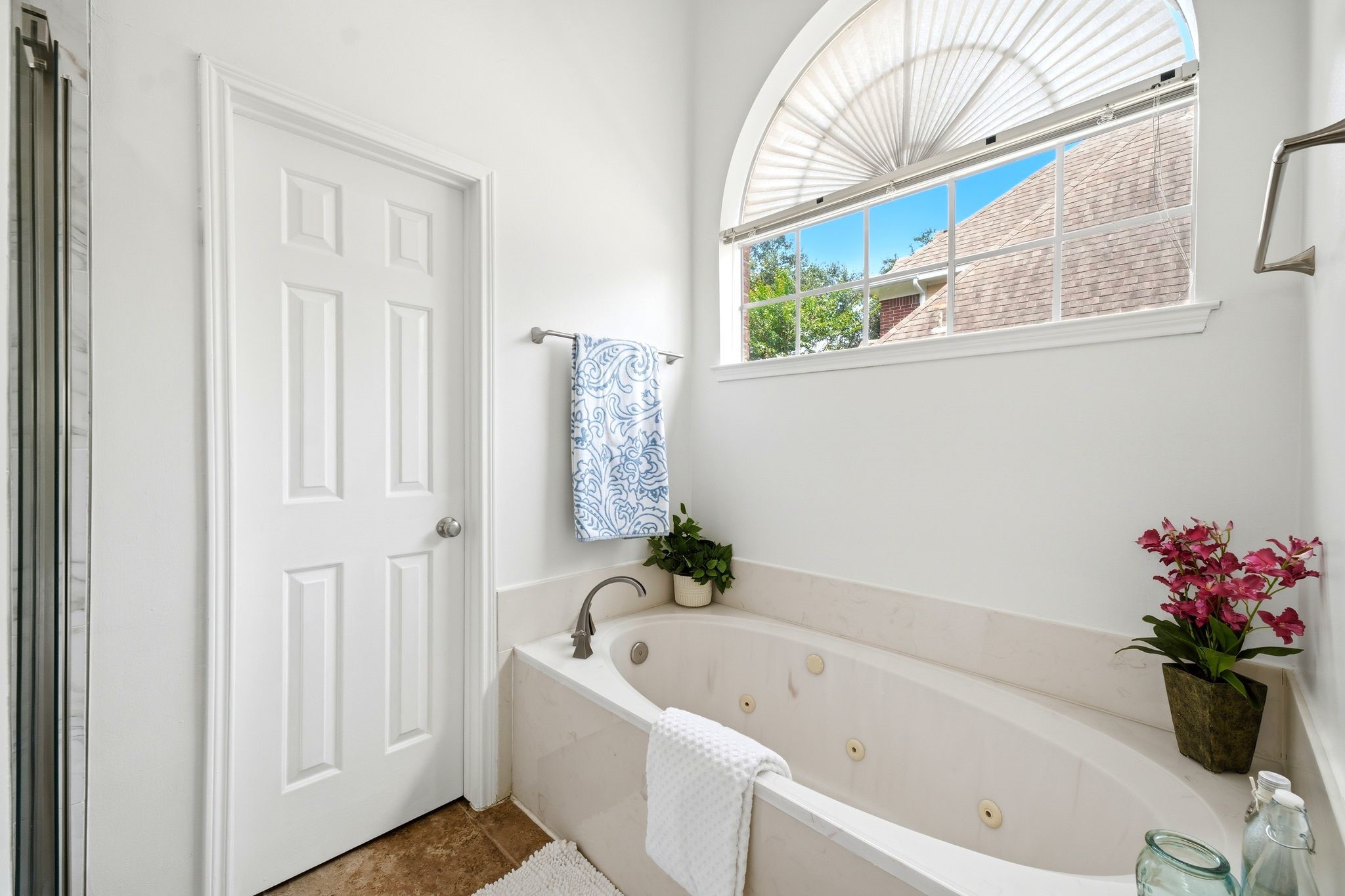 Experience serene luxury in this bright bathroom, featuring a jetted bathtub beneath an arched window, enhanced by elegant fixtures and lush decorative plants.