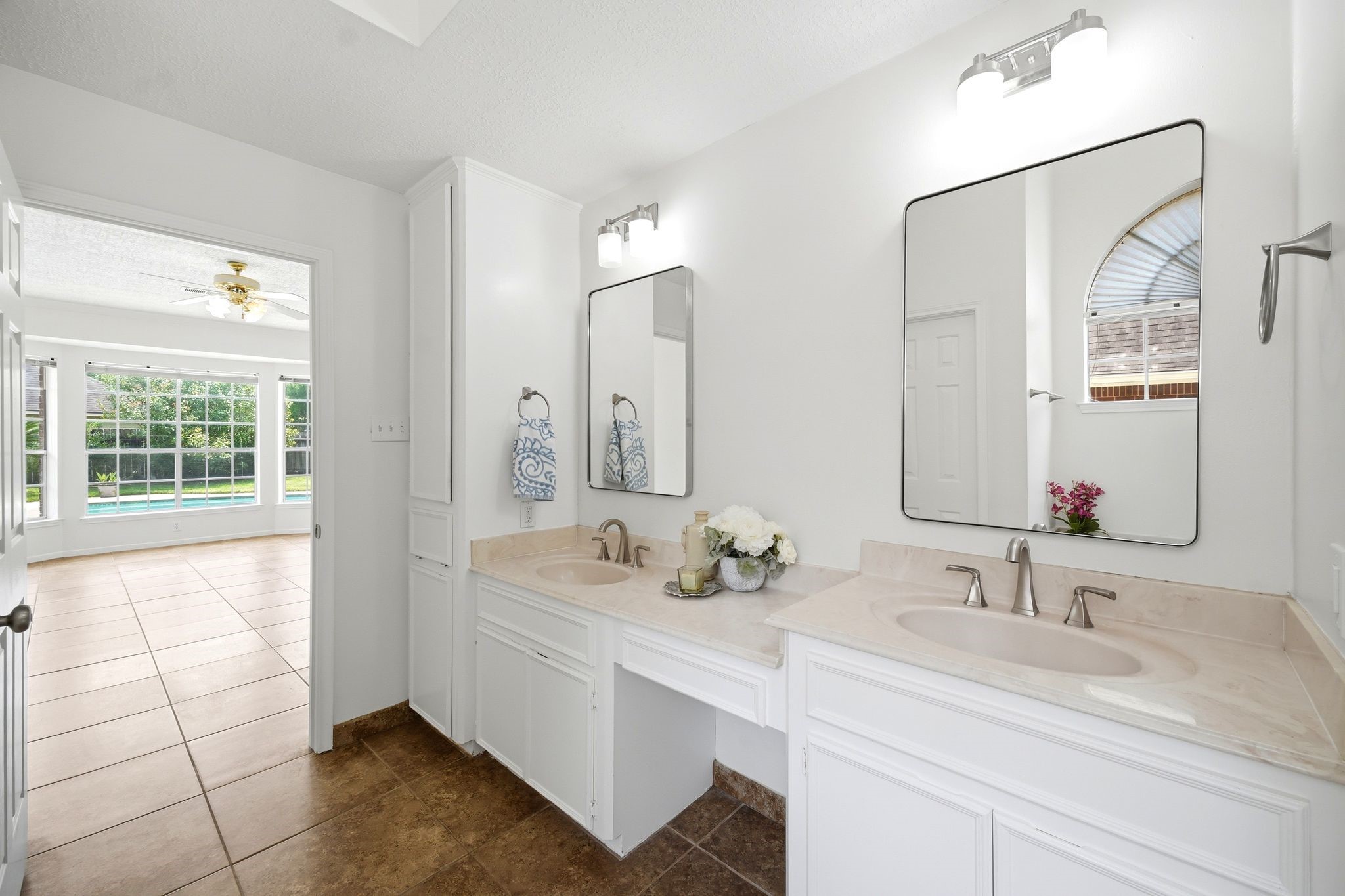 Experience luxury in this bright, spacious bathroom with dual sinks, modern lighting, and expansive mirrors, leading into a sunlit room with tiled flooring and a large window showcasing a pool view.