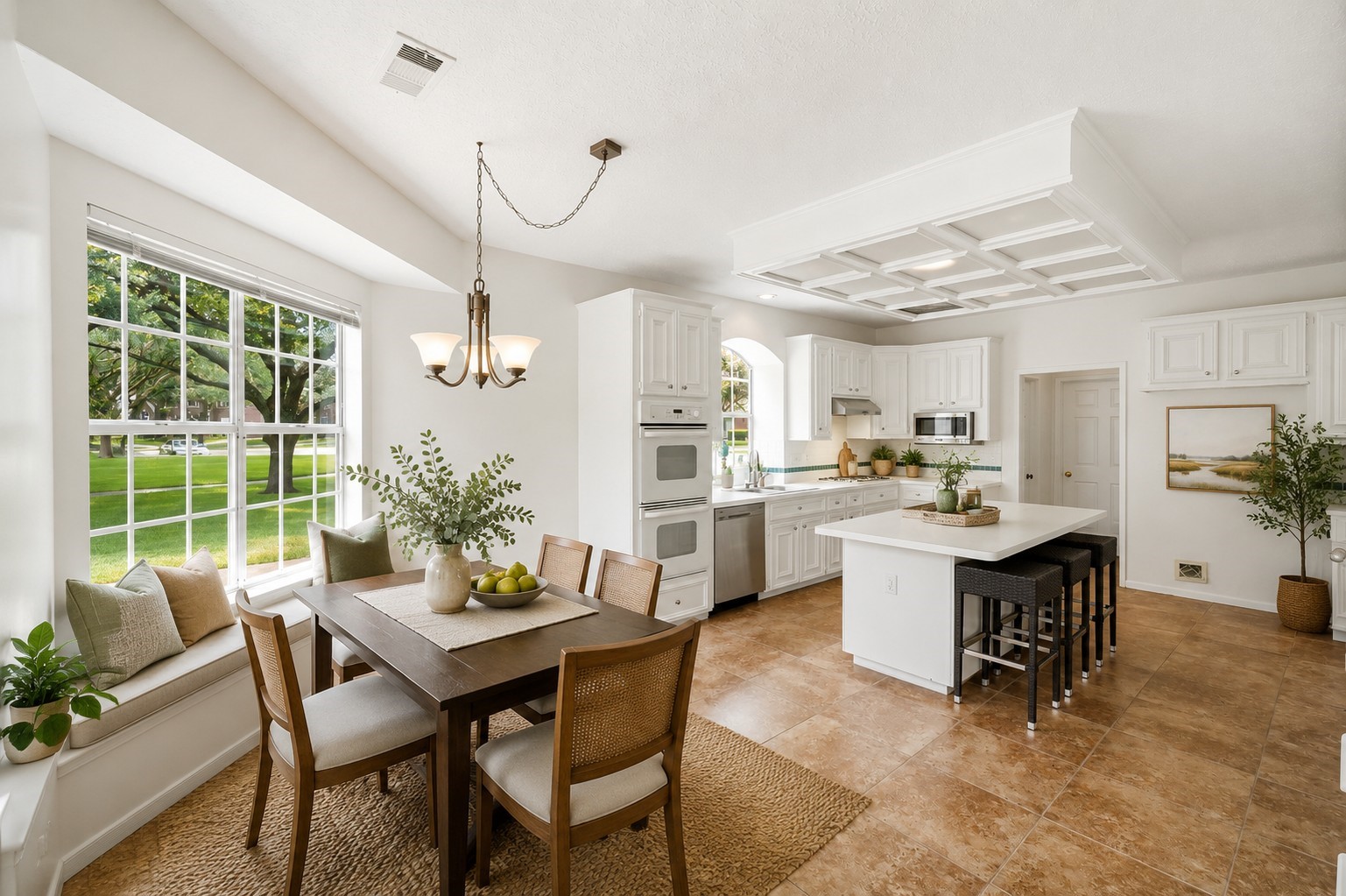 Experience the perfect blend of style and comfort in this bright kitchen, featuring elegant white cabinetry, a central island with bar seating, and a cozy dining nook with large windows offering serene views of a lush green yard.