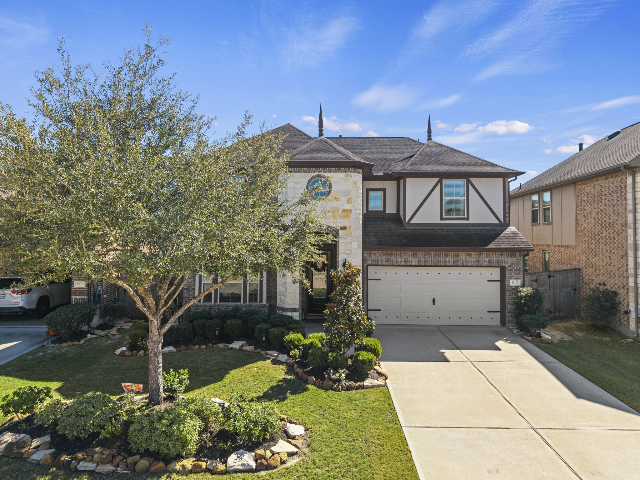 Charming two-story home with a spacious driveway, elegant brick and stone exterior, and well-manicured landscaping featuring mature trees and shrubs.