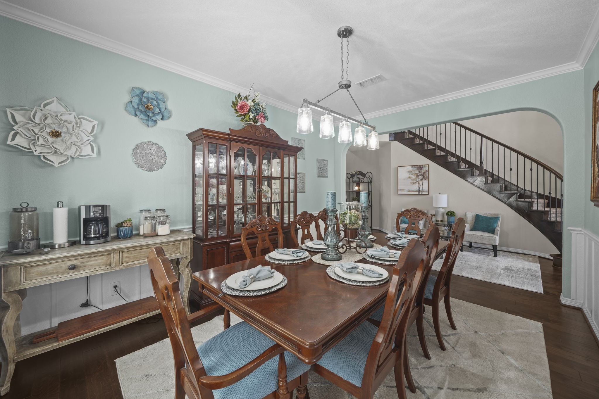 Elegant dining room with a grand wooden table, stylish chairs, and a striking chandelier. The space is accented by a beautiful china cabinet and decorative wall art, leading to an open staircase for a touch of sophistication.