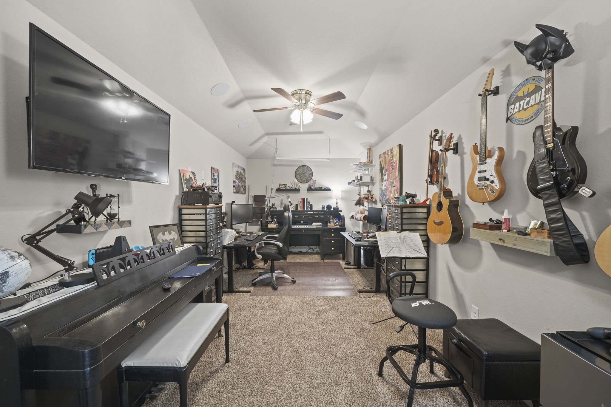 Versatile music and workspace featuring wall-mounted guitars, a digital piano, ample shelving for storage, and a large TV, all under a ceiling fan for comfort.