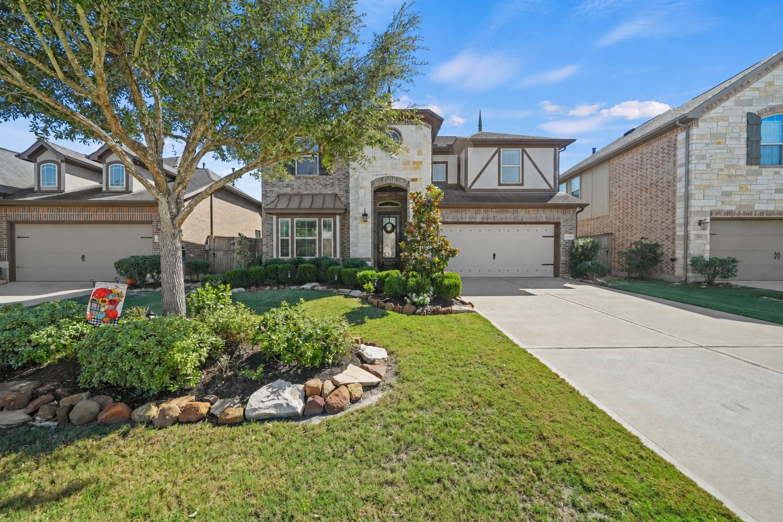 Charming two-story home with a stone facade and a welcoming front porch, nestled in a beautifully landscaped yard with mature trees and manicured shrubs.