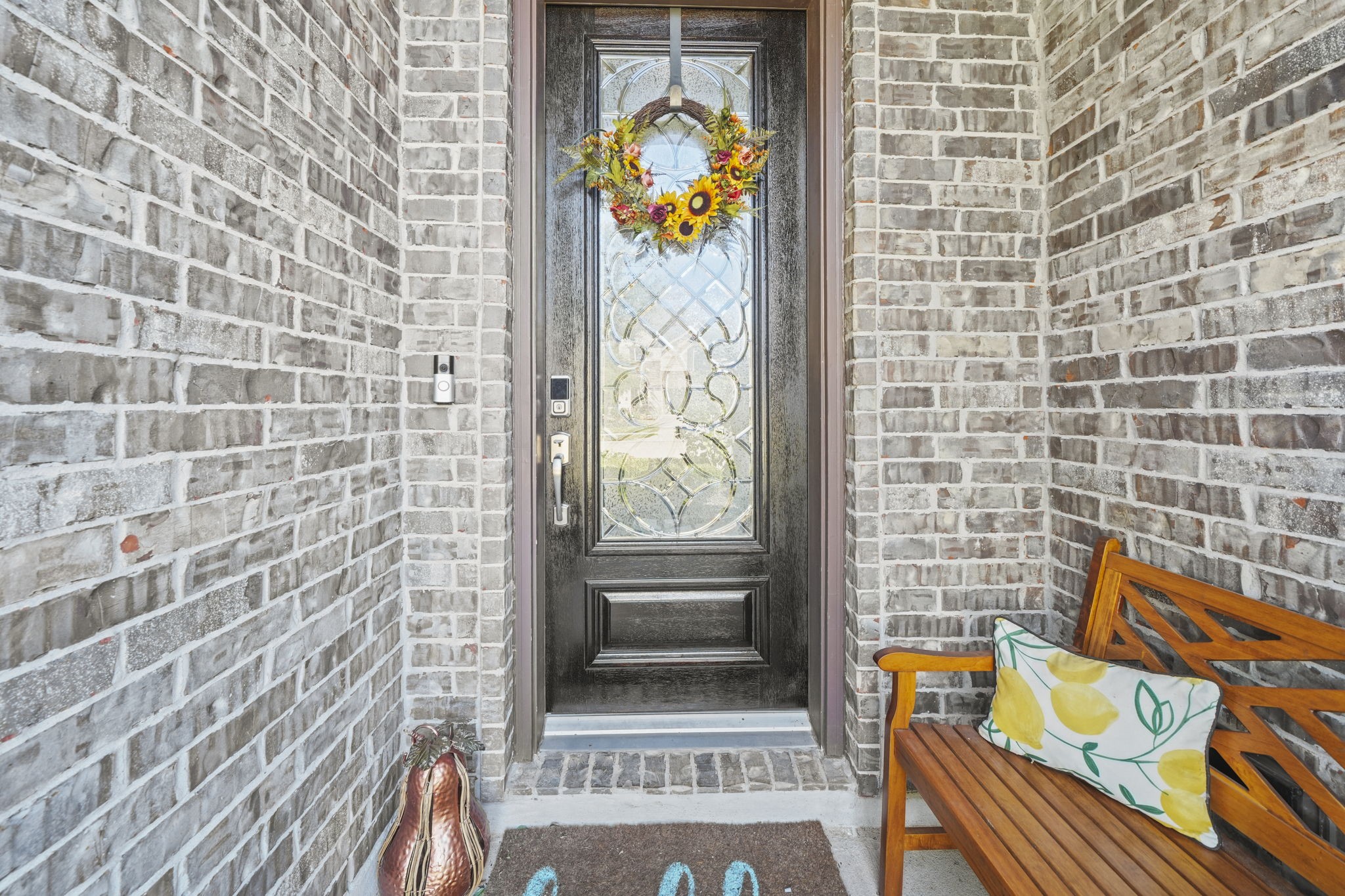 Charming brick entryway with a decorative glass front door, adorned with a vibrant floral wreath. Cozy bench seating adds a welcoming touch.