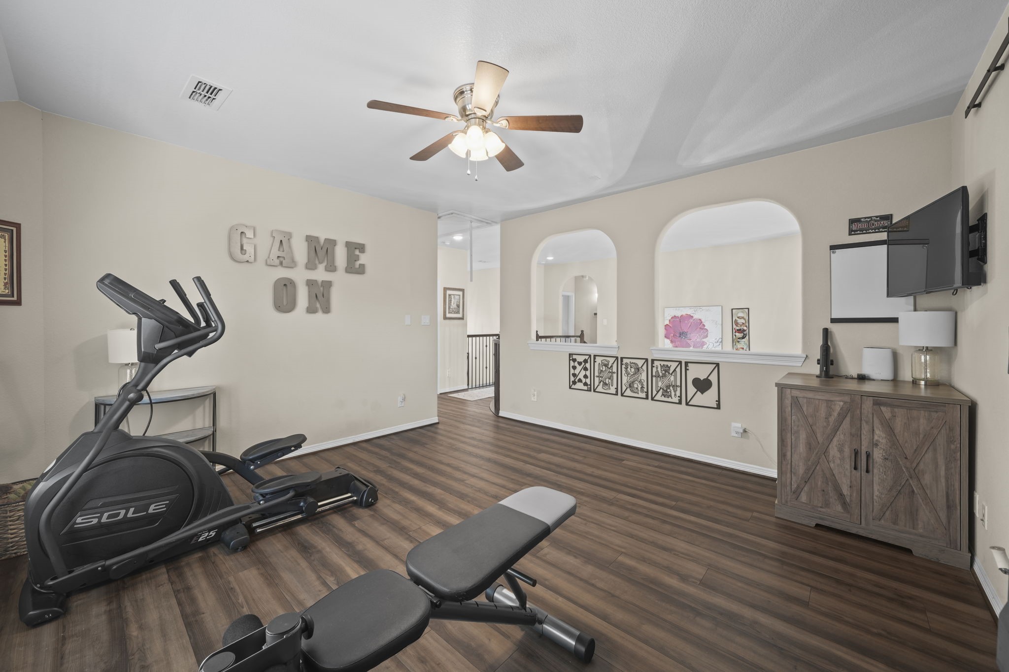 Spacious room with warm wood flooring, ideal for a home gym or entertainment area, featuring a ceiling fan, exercise equipment, and stylish decor.