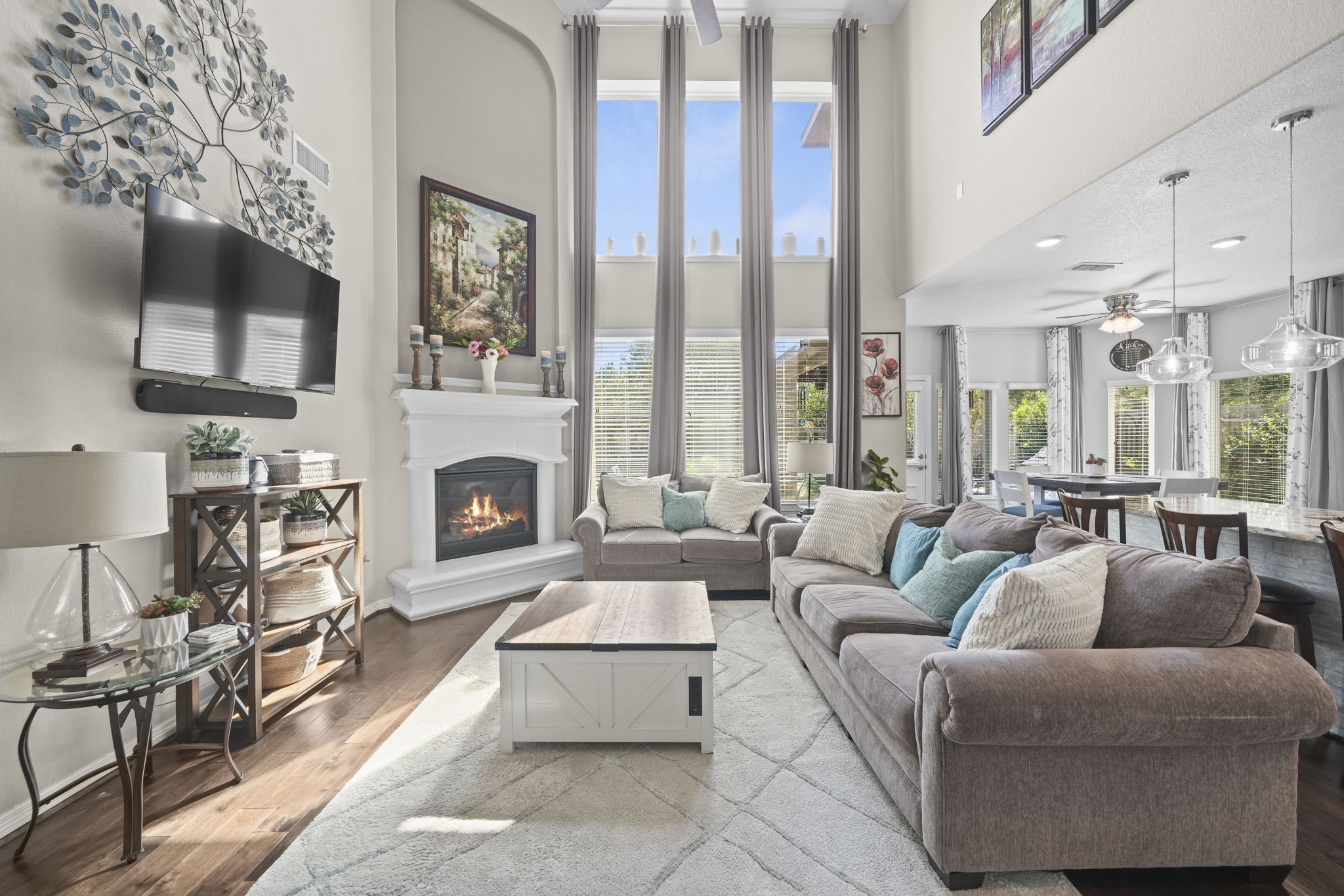 Spacious and elegant living area with soaring ceilings and abundant natural light, featuring a cozy fireplace, modern decor, and an open layout connecting to the dining area and kitchen.