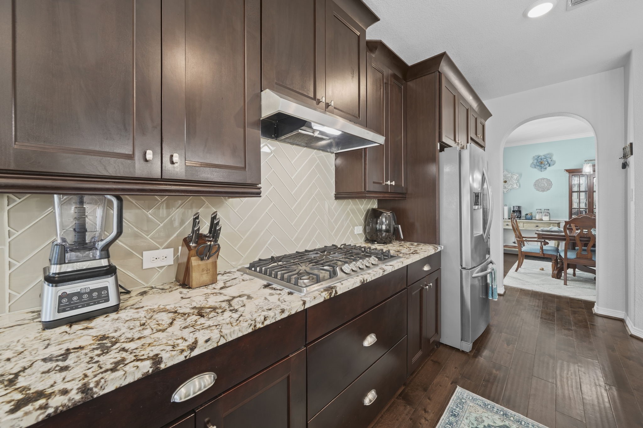 Elegant kitchen with rich dark wood cabinets, sleek stainless steel appliances, and a stylish herringbone tile backsplash. The granite countertops provide ample workspace, while the open archway leads to a cozy dining area.