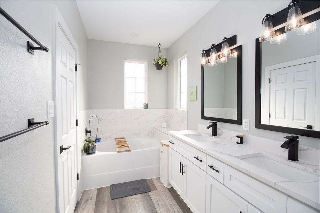 Relax and unwind in this beautifully appointed primary bathroom featuring a soaking tub, elegant marble-look tile accents, white cabinetry, stylish black fixtures, and natural light from two windows.