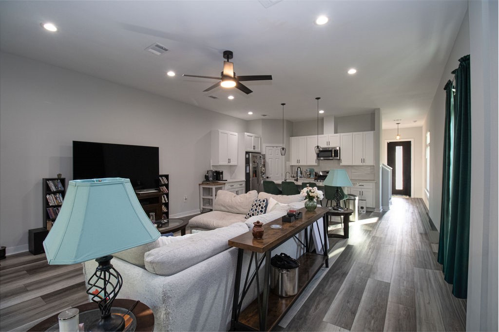 Bright and welcoming living area featuring multiple windows, attractive wood-look flooring, a modern ceiling fan, and plenty of room for comfortable seating and entertaining.