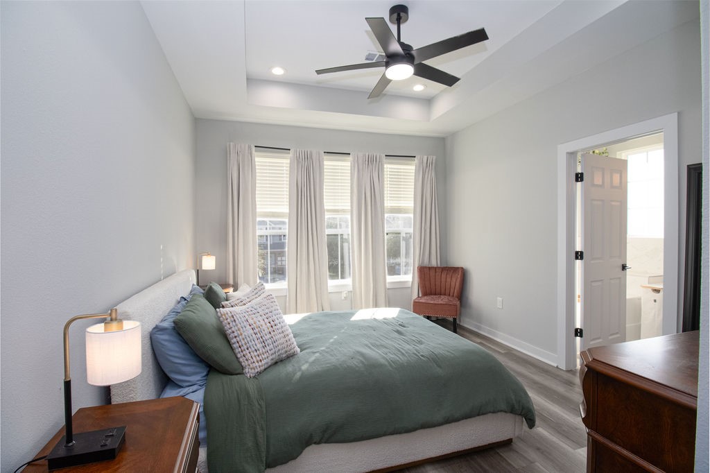 Spacious and inviting primary retreat featuring a decorative tray ceiling, modern ceiling fan, beautiful wood-look flooring, and three windows that fill the room with natural light.