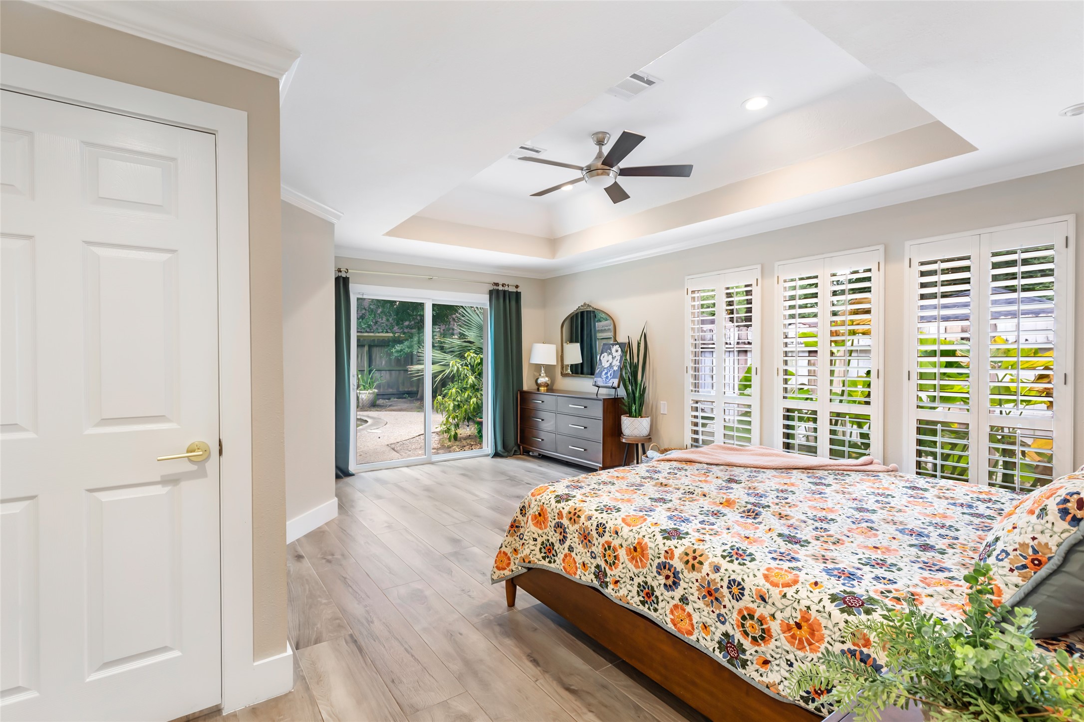 Spacious and bright primary bedroom featuring elegant tray ceilings and a modern ceiling fan, with large windows and sliding glass doors leading to a lush outdoor area, offering an inviting and serene retreat.