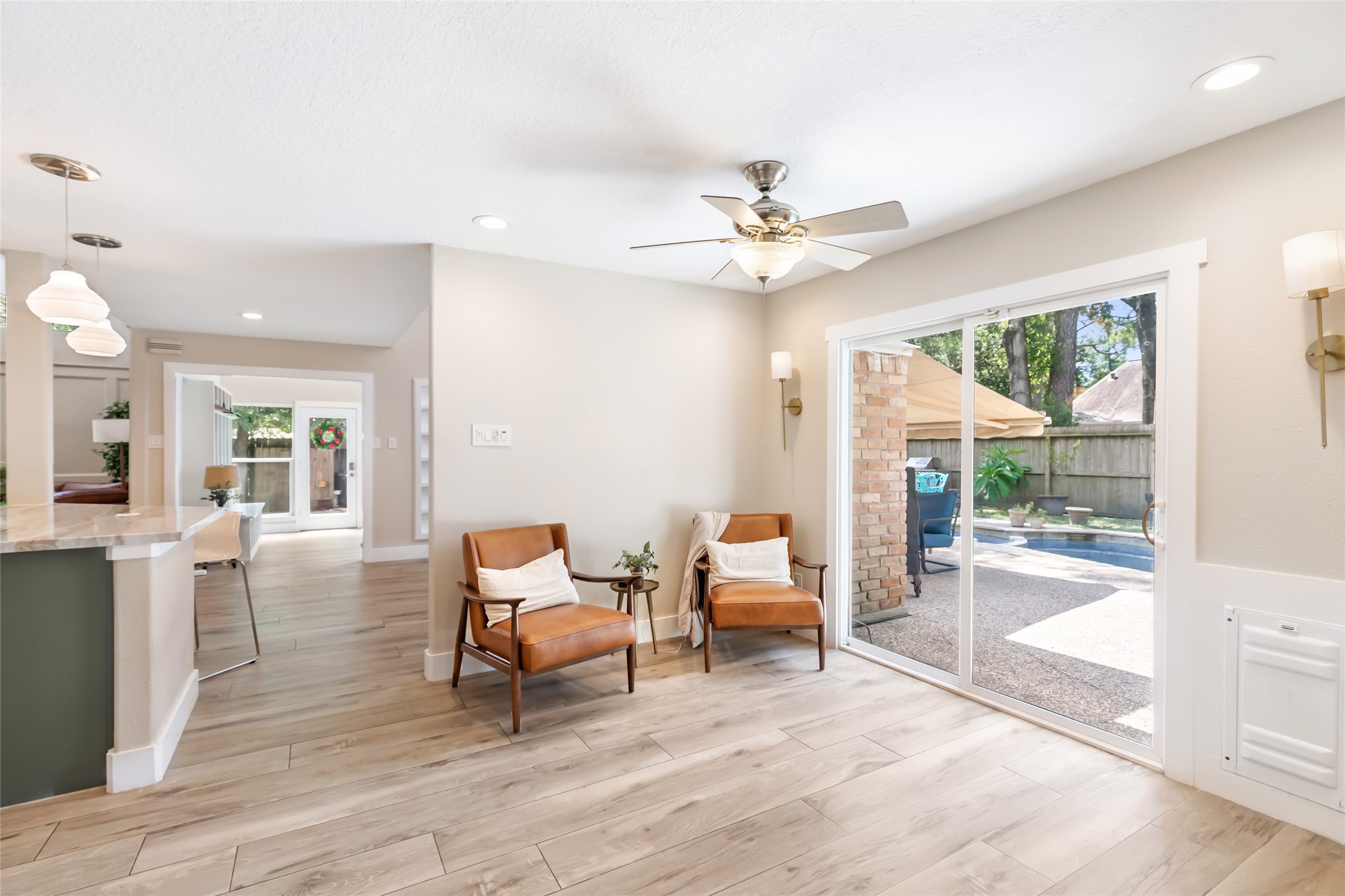 Bright and inviting versatile living space with stylish wood flooring, cozy seating, and large sliding glass doors leading to a charming outdoor patio and pool area.