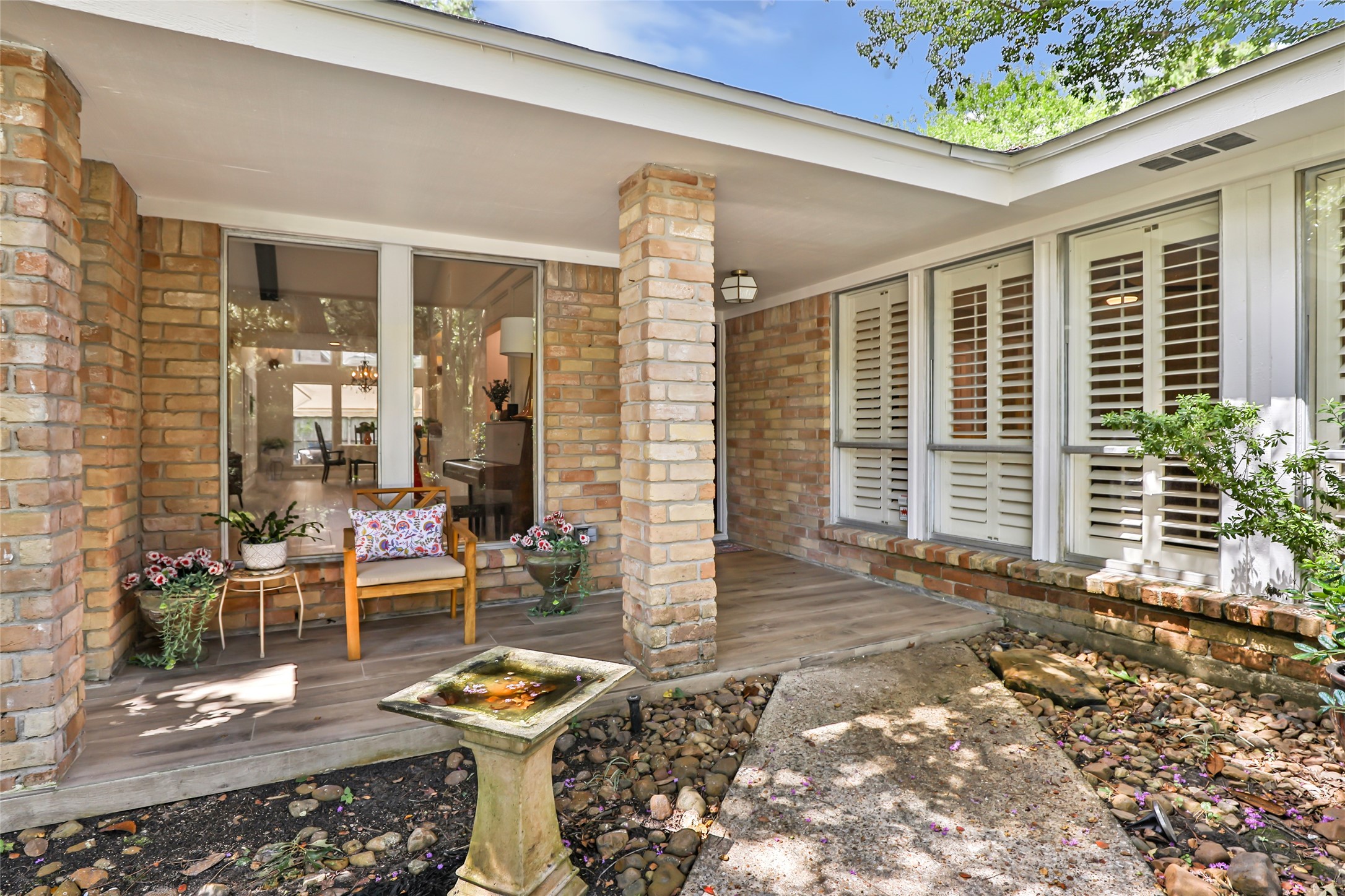 Charming front porch with elegant brick columns, large windows with plantation shutters, and a cozy sitting area surrounded by lush greenery and a decorative birdbath. Charming front porch with elegant brick columns, large windows with plantation shutters, and a cozy sitting area surrounded by lush greenery and a decorative birdbath.