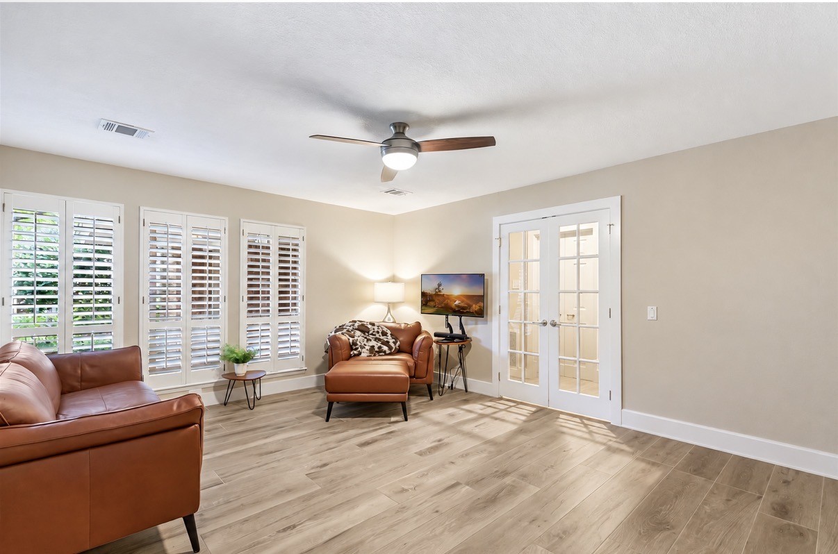 Bright and inviting living space featuring elegant ceramic wood flooring, comfortable seating, and ample natural light through plantation shutters. A versatile area perfect for relaxation or a home office setup.