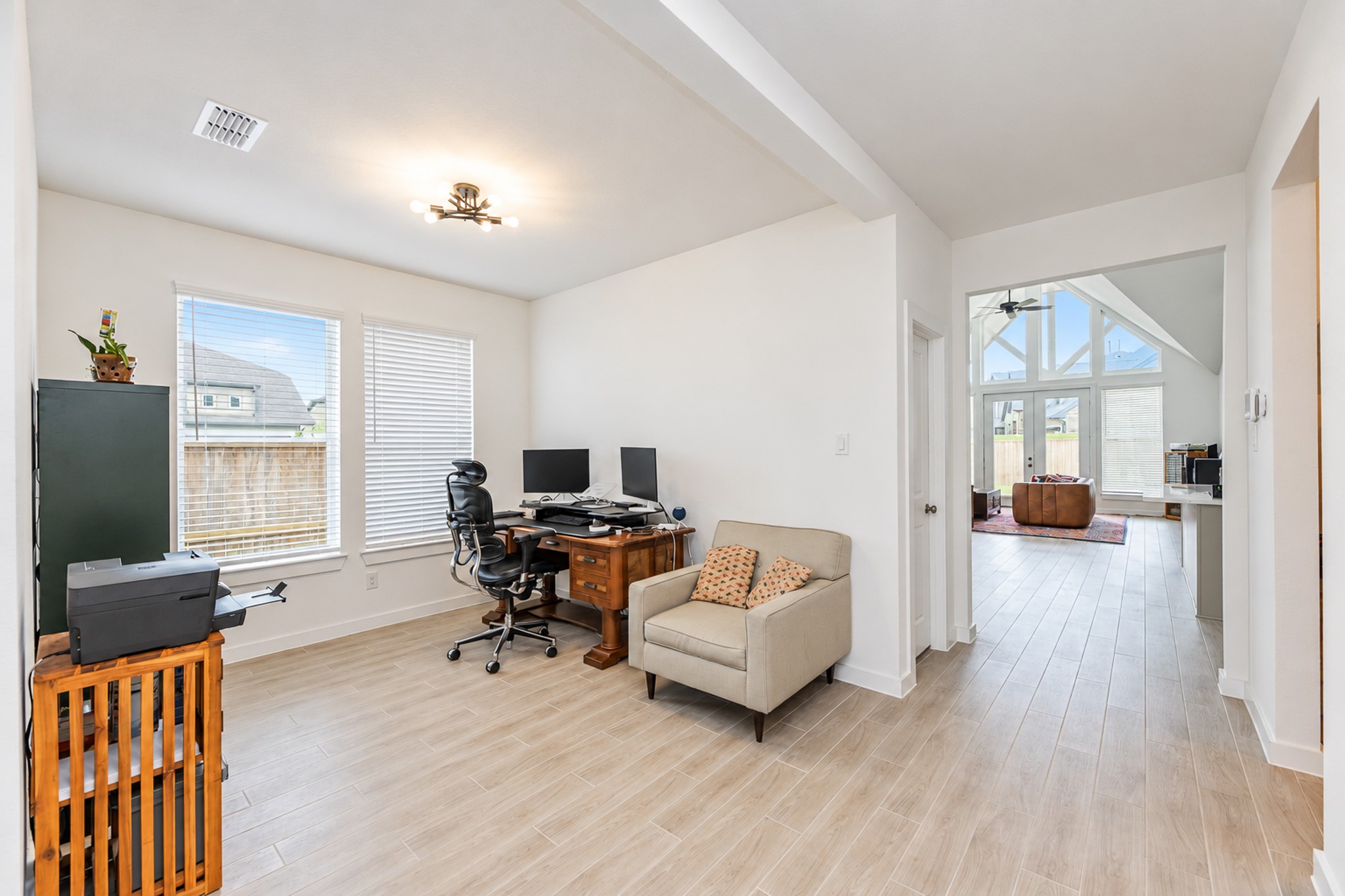 Bright and inviting home office with elegant wood flooring, ample natural light from large windows, and an open view leading into a spacious living area with vaulted ceilings.