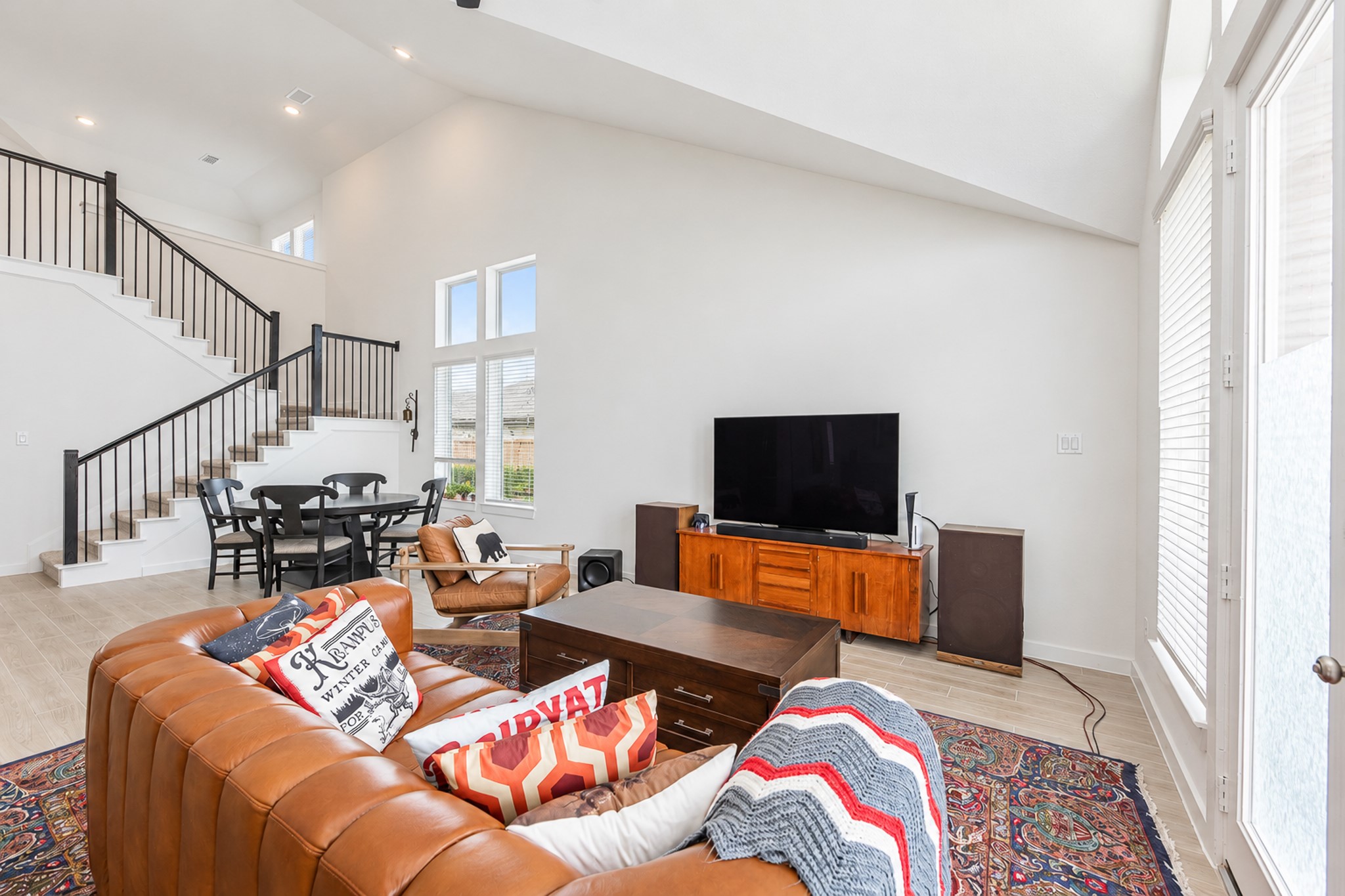 Spacious living area with high ceilings, featuring a cozy leather sofa, a large TV, and natural light from tall windows. The open staircase leads to a charming dining nook, enhancing the inviting atmosphere.