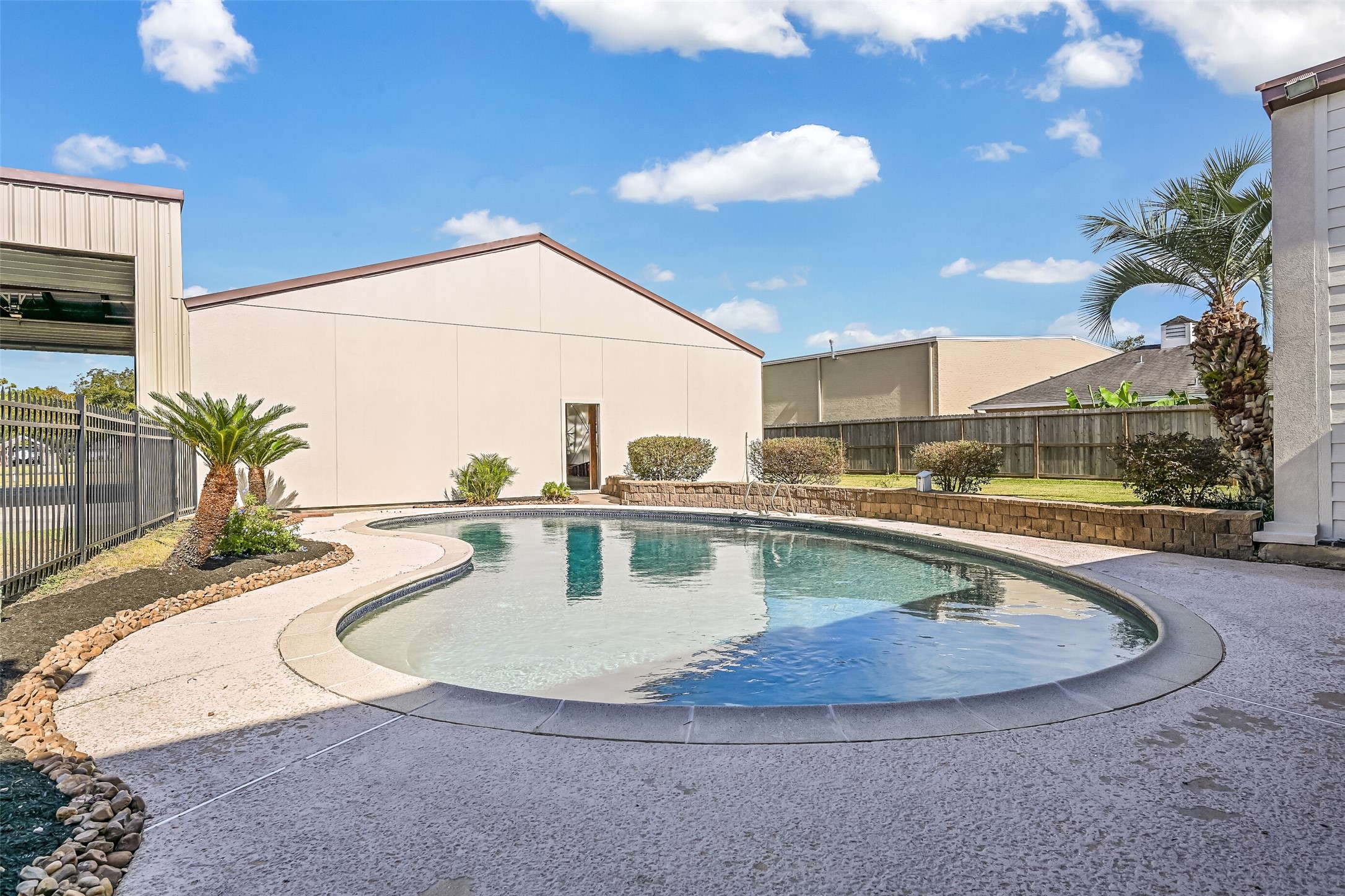 The backyard swimming pool features a graceful freeform design surrounded by textured concrete decking, tropical landscaping & decorative stone borders. A raised planting bed adds dimension to the space, while the adjoining covered patio provides a shaded retreat just steps from the water.