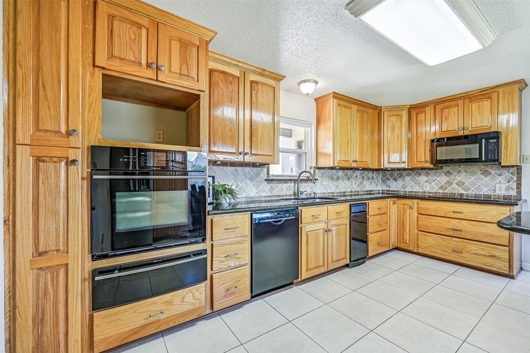 Generous counter space & extensive wood cabinetry line this functional kitchen, complete with granite countertops, a decorative tile backsplash, double-basin sink & black appliances. Built-in wall ovens, a separate cooktop & multiple drawers provide ample space for meal preparation and storage.