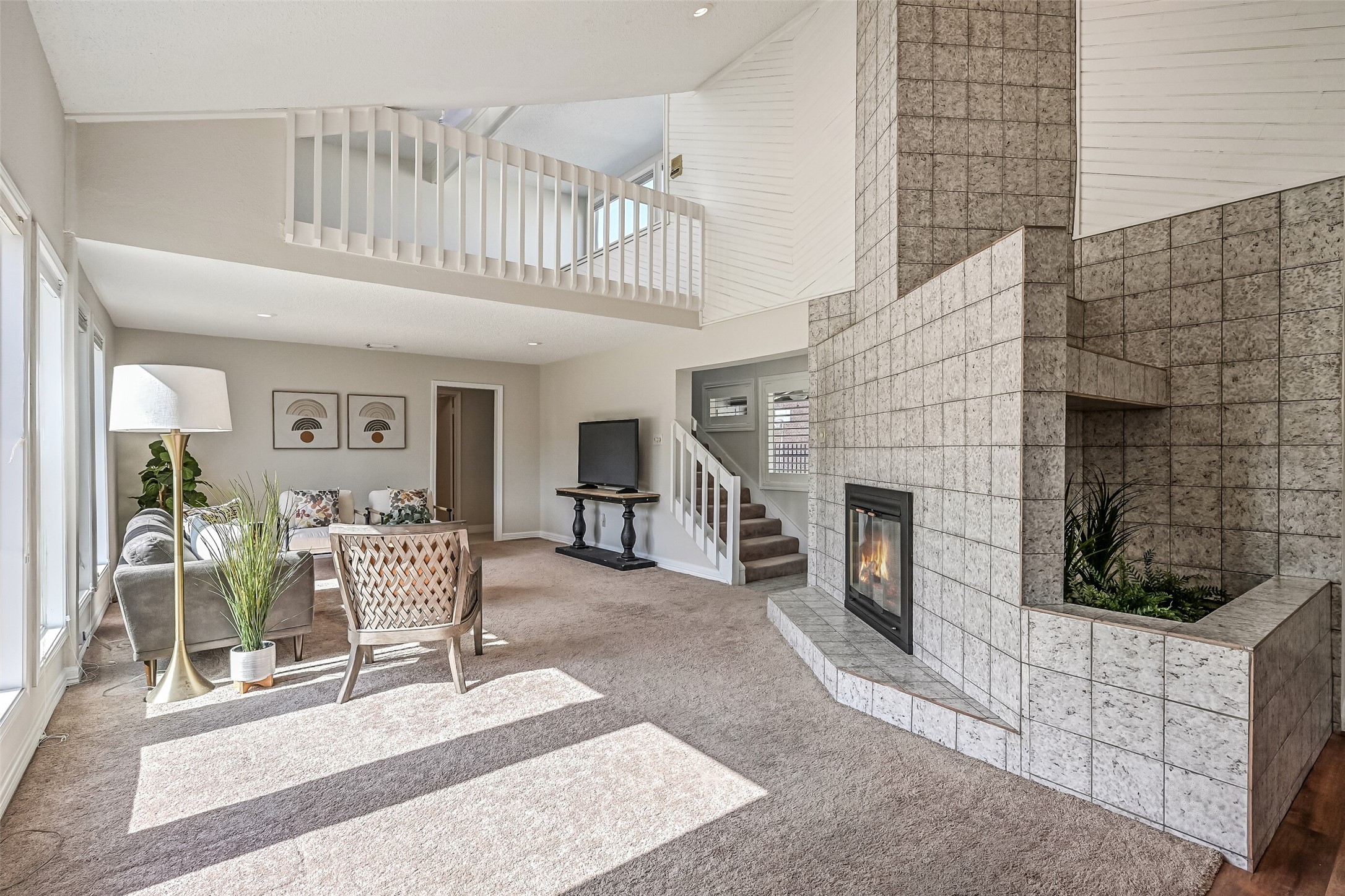 The soaring living room is anchored by a striking floor-to-ceiling tiled fireplace with an adjoining decorative planter. Natural light streams through tall windows, while white architectural detailing & an open loft above add dimension to this inviting gathering space.