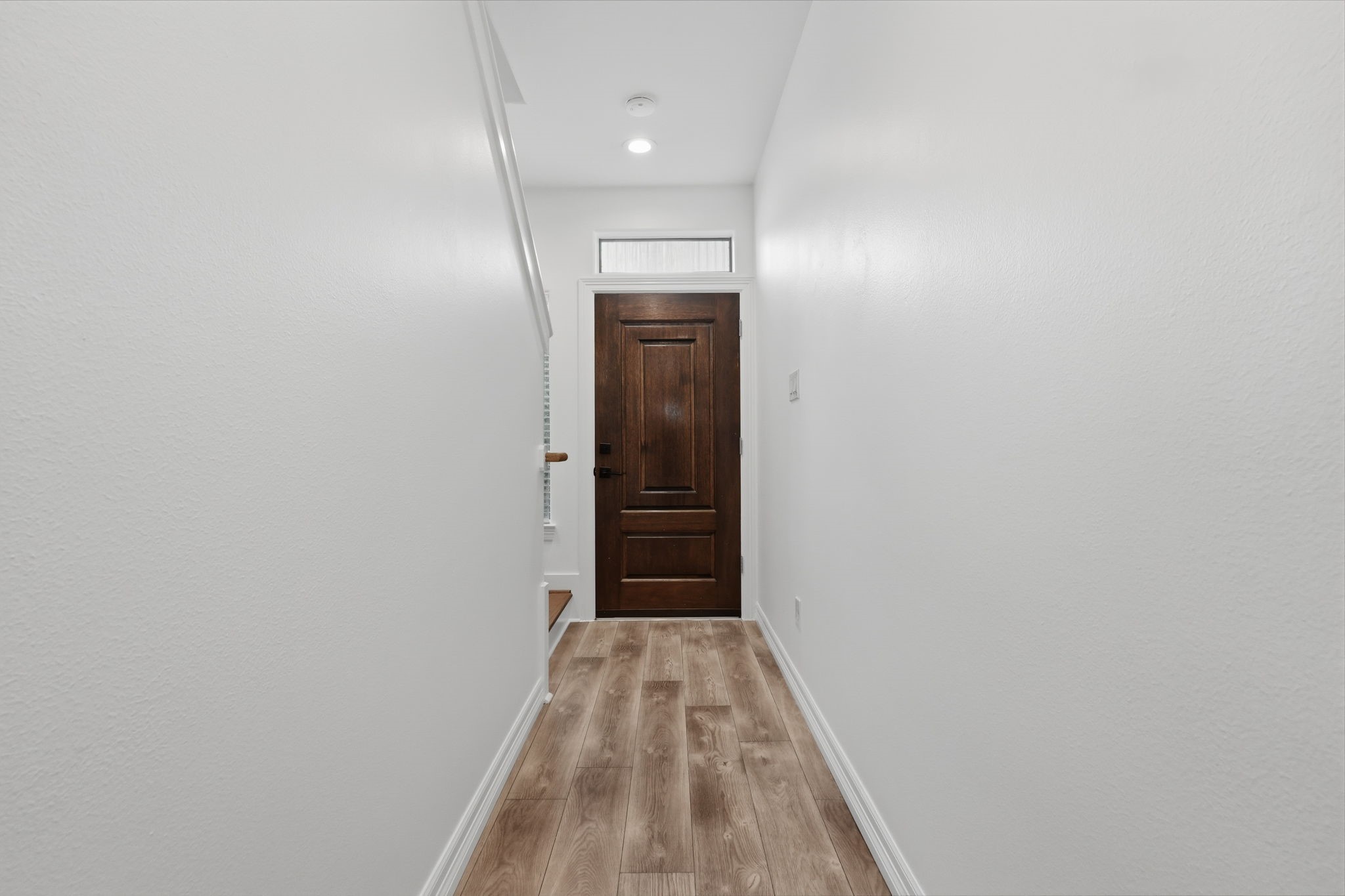 The hallway features crisp white walls, recessed ceiling lighting, and a rich wood door at the end, creating a clean, streamlined transition between living spaces.