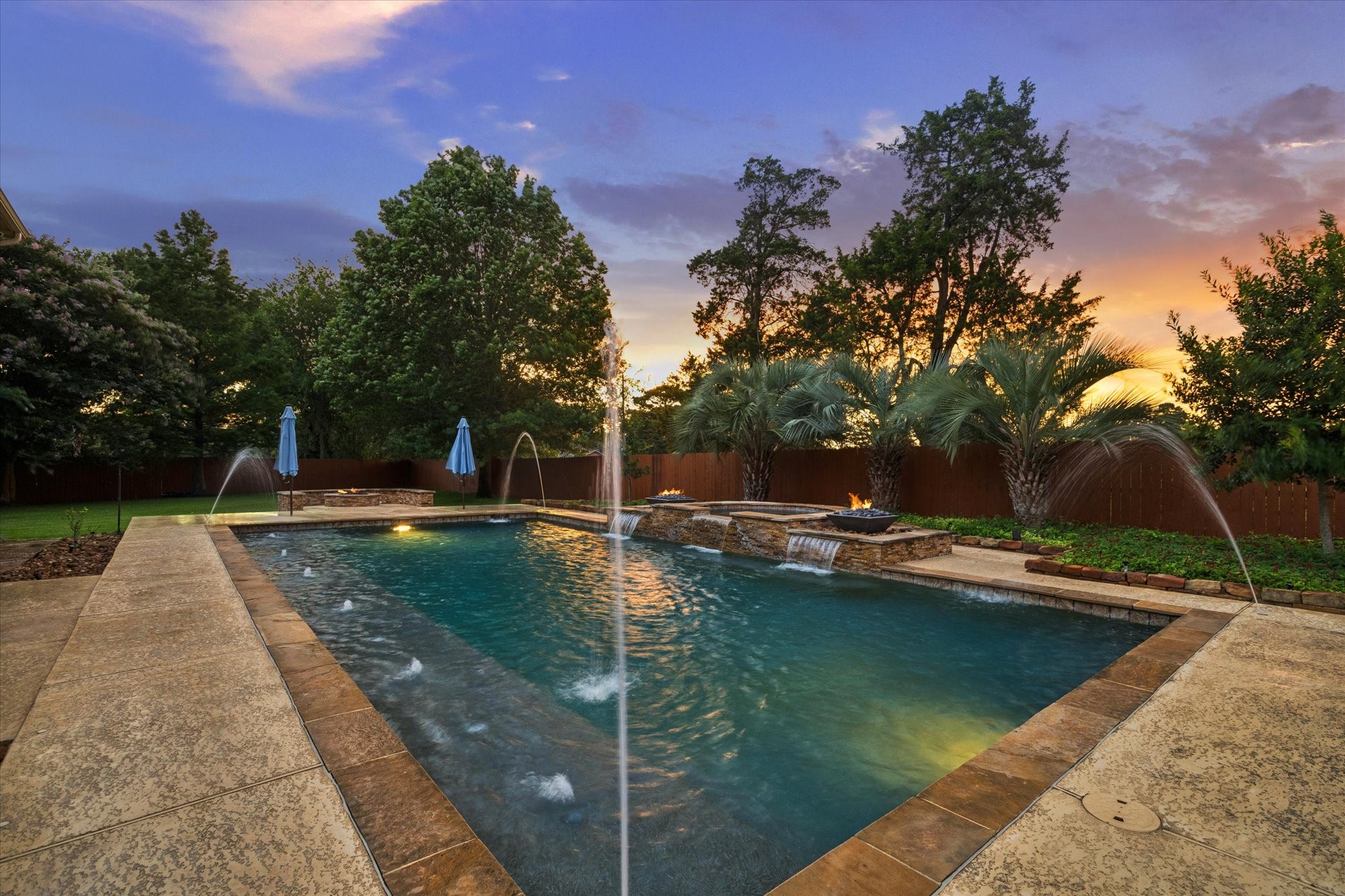 A stunning poolside retreat surrounded by mature trees and lush landscaping, where dramatic water features and glowing fire accents create a serene and inviting atmosphere at sunset.