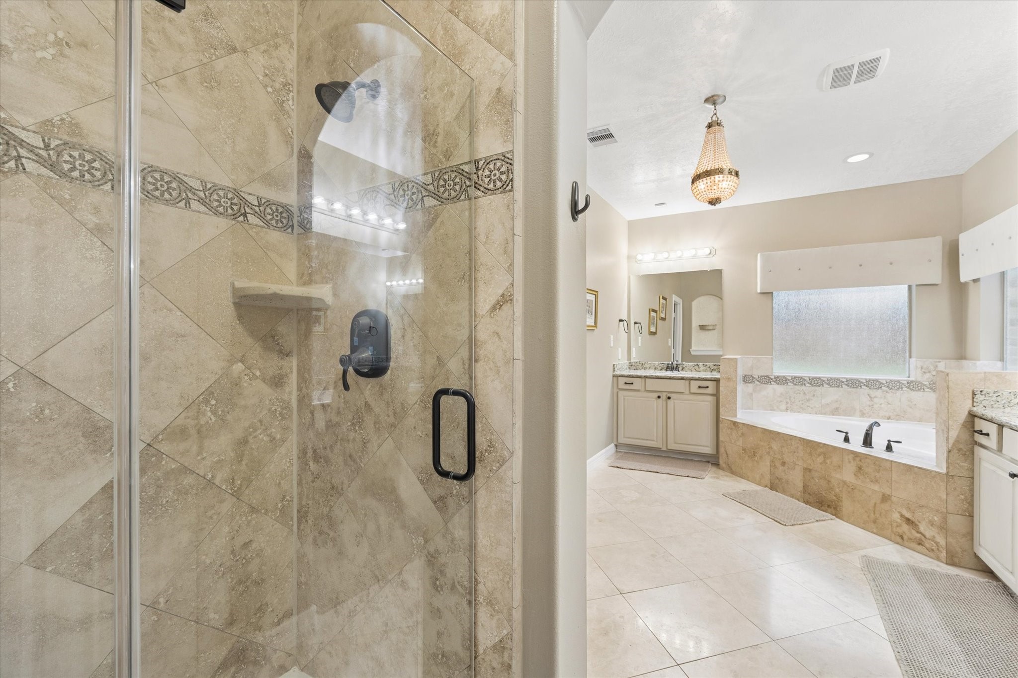 A closer view of the generously sized primary en-suite bath highlights the separate walk-in shower with decorative tile accents and glass enclosure, complemented by the jetted soaking tub and dual vanities. Abundant natural light enhances the open, airy atmosphere, creating a relaxing and functional private retreat.