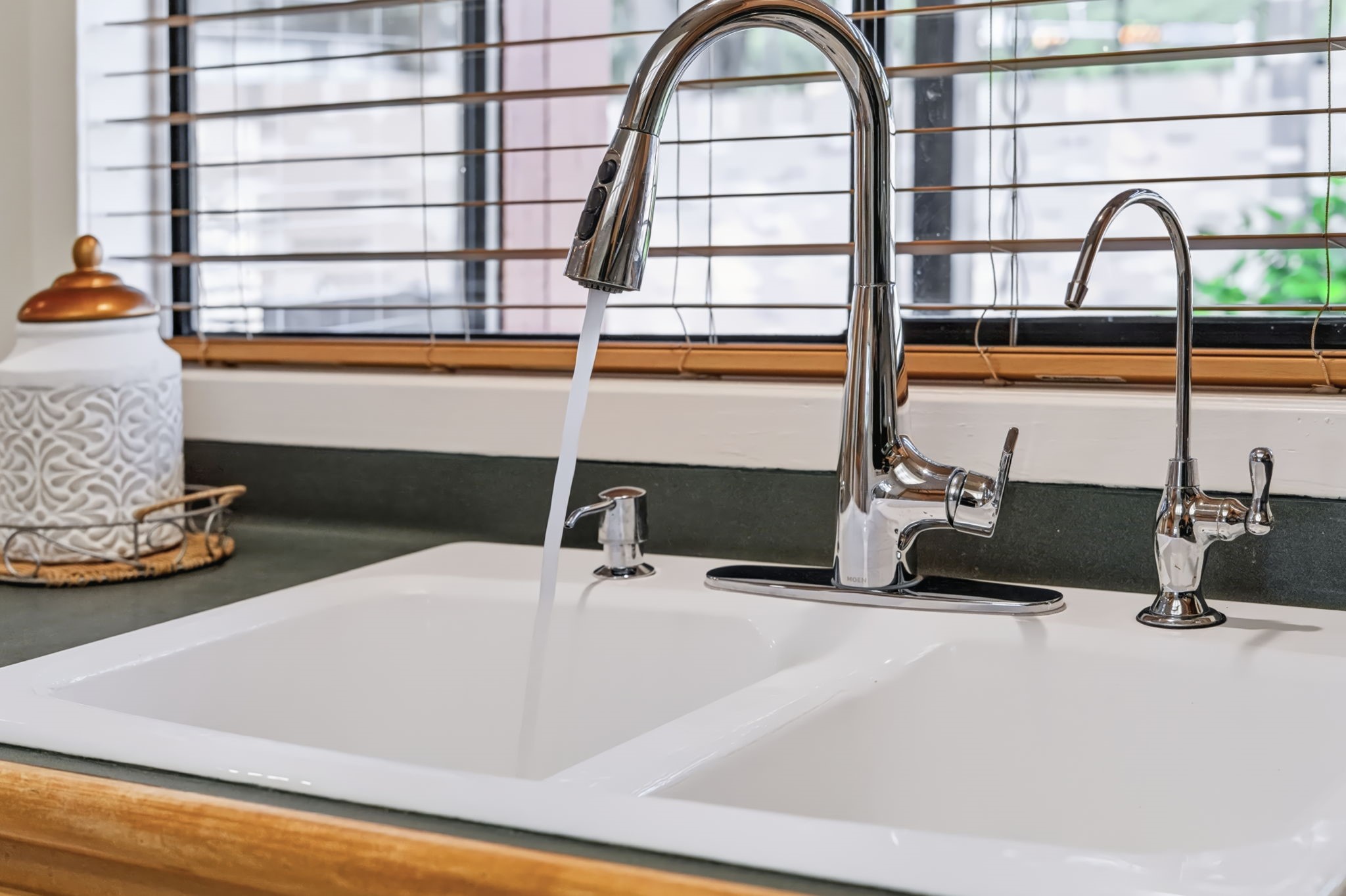 A double porcelain sink features a gooseneck pull-down faucet and a separate water filtration faucet, while the window above offers a relaxing view of the backyard.