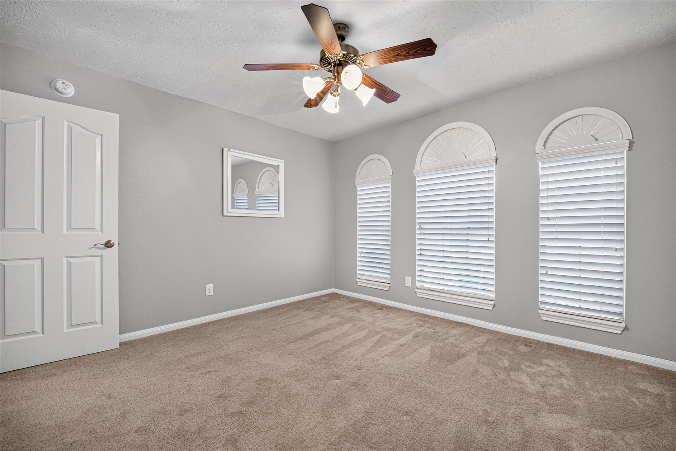 Secondary bedroom features three arched windows with white blinds, a ceiling fan, carpeted flooring, and a paneled entry door.