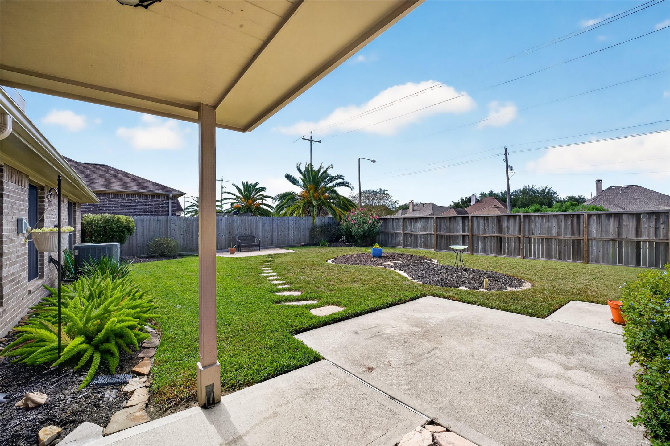 Covered rear patio overlooks the fenced backyard, with concrete paving, planting beds, and a stepping-stone path extending into the lawn.