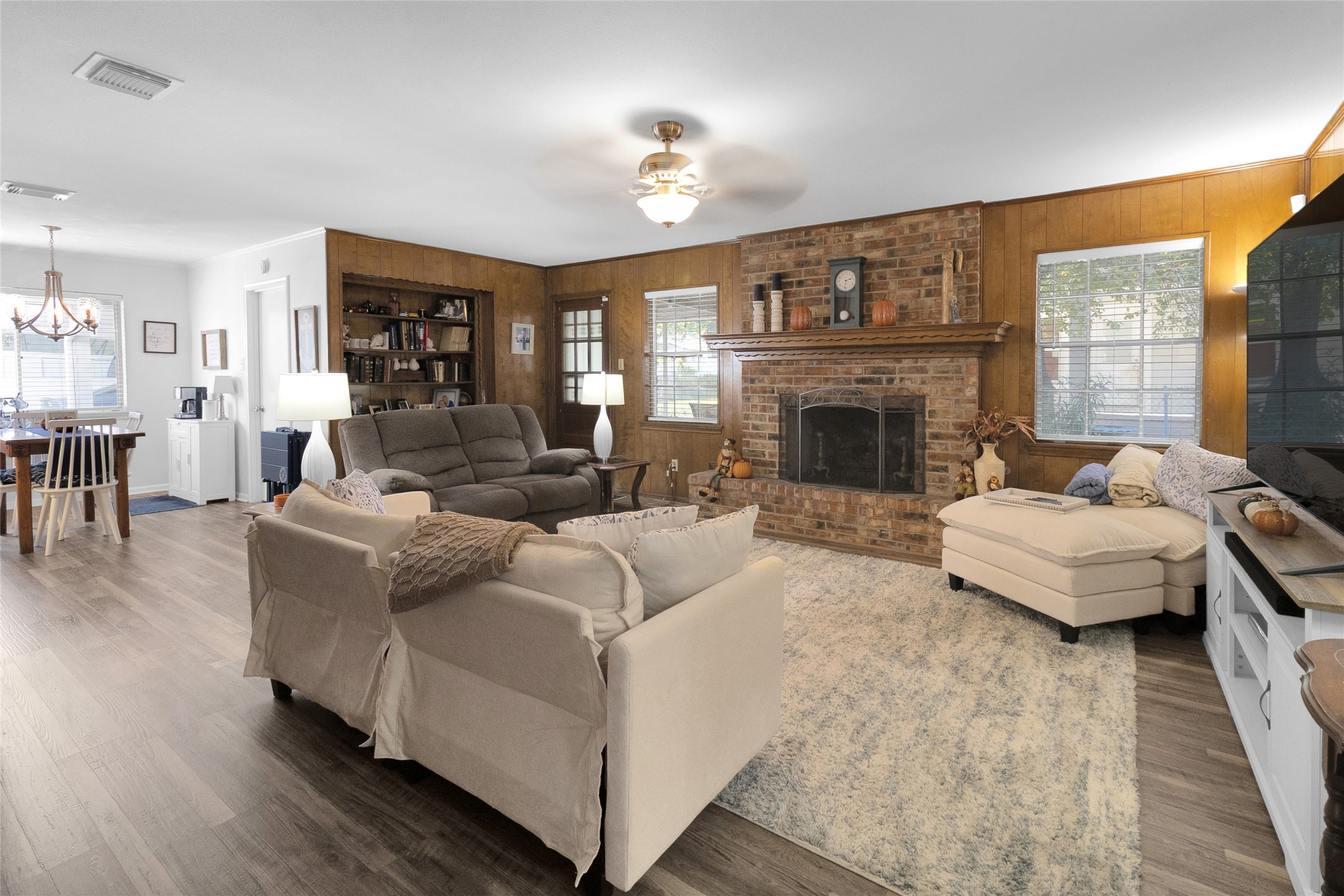 Charming living room with a cozy brick fireplace, rich wood paneling, and ample natural light. The open layout seamlessly connects to the breakfast area, offering an inviting space for relaxation and entertaining. Charming living room with a cozy brick fireplace, rich wood paneling, and ample natural light. The open layout seamlessly connects to the breakfast area, offering an inviting space for relaxation and entertaining.