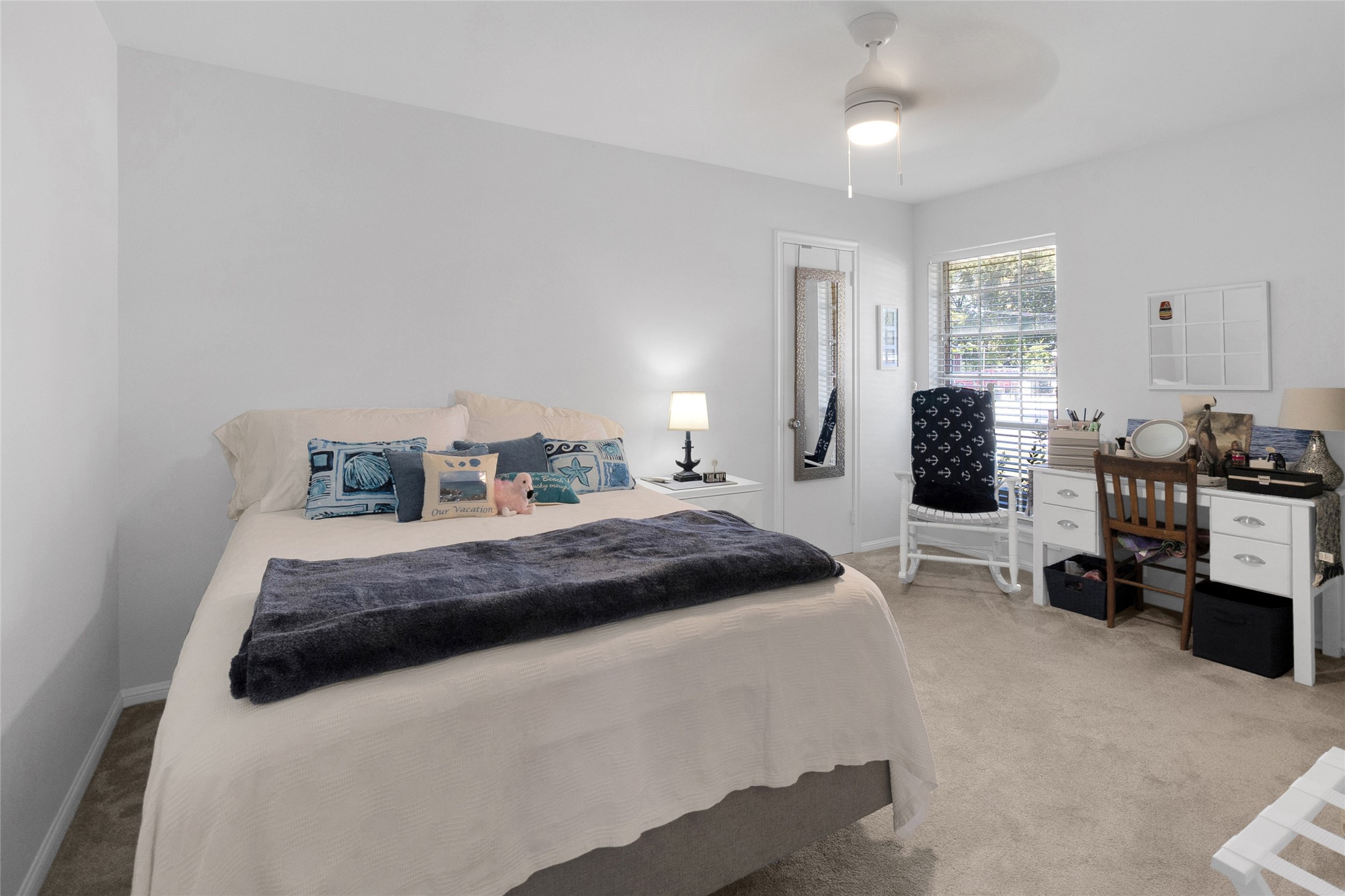 Second bedroom with soft carpeting, a comfortable bed adorned with decorative pillows, and ample natural light from a large window. A ceiling fan and a workspace add functionality, while neutral tones create a serene atmosphere.
