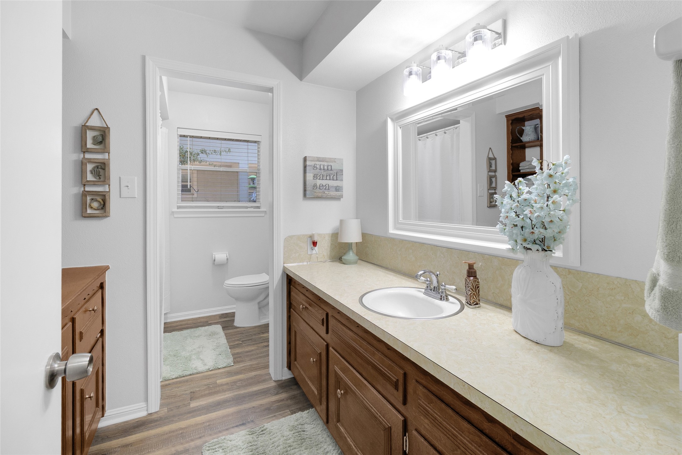 Charming bathroom with a large mirror, ample countertop space, and warm wood cabinetry. The room is brightened by soft lighting and complemented by tasteful decor, creating a welcoming and functional space.