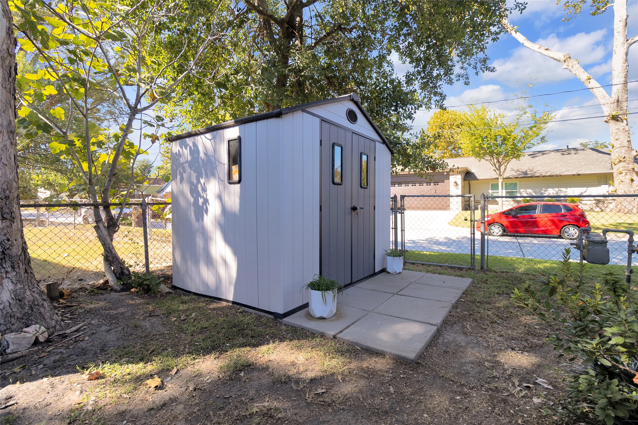 A charming, well-maintained garden shed sits in a fenced backyard, surrounded by mature trees and enhanced by a paved pathway, offering functional storage space.