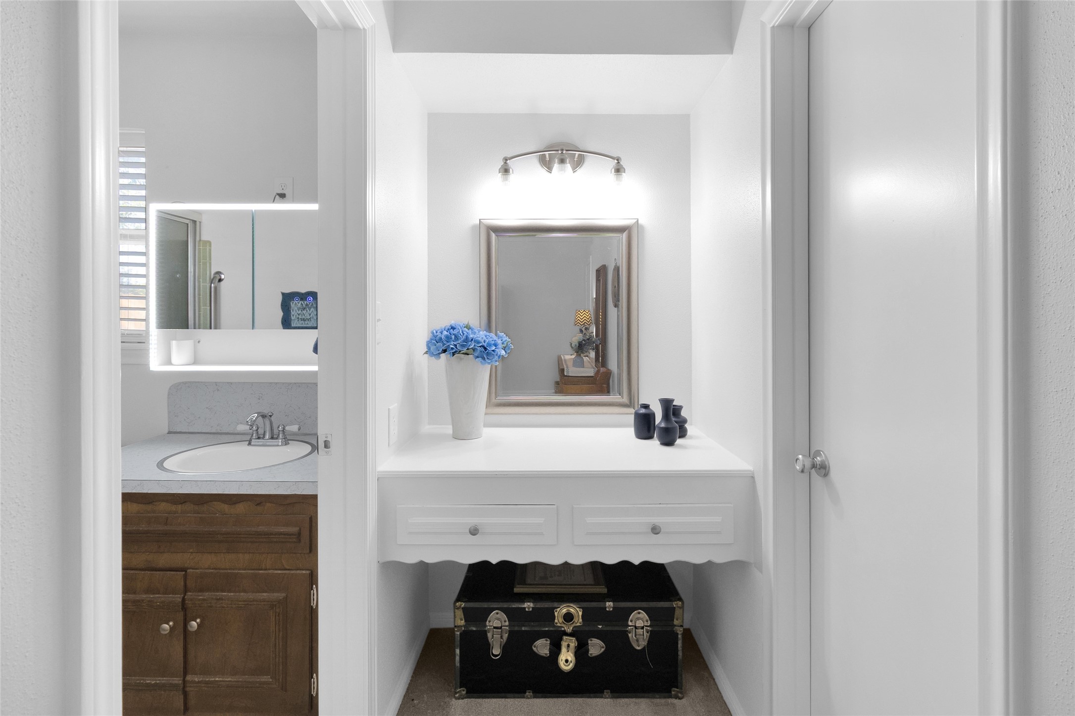 Charming vanity area with elegant lighting and a built-in mirror, adjacent to a well-appointed bathroom featuring a stylish sink and wood cabinetry.
